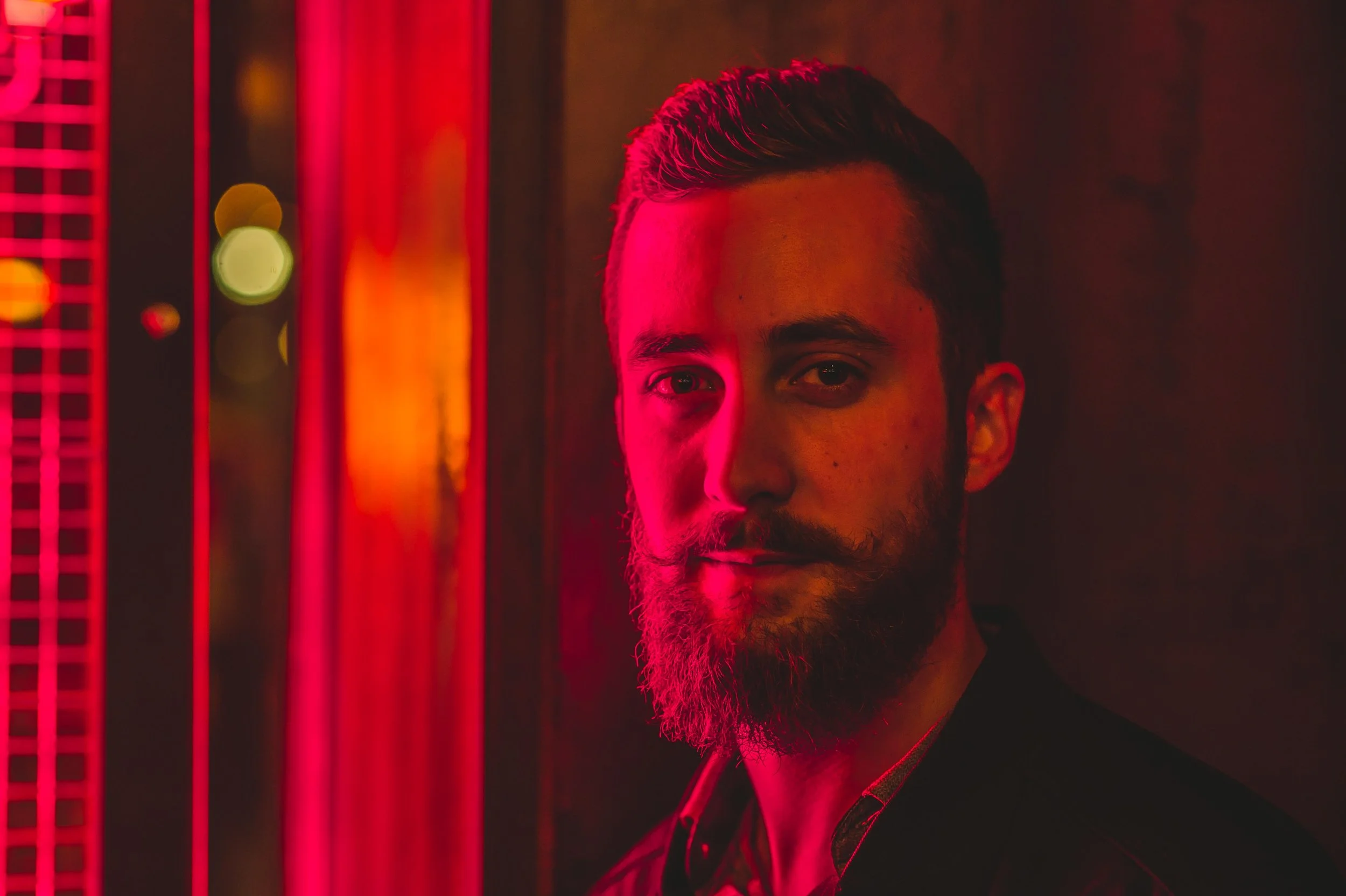 A man with a beard and mustache in a dark room with red lighting, looking at the camera.