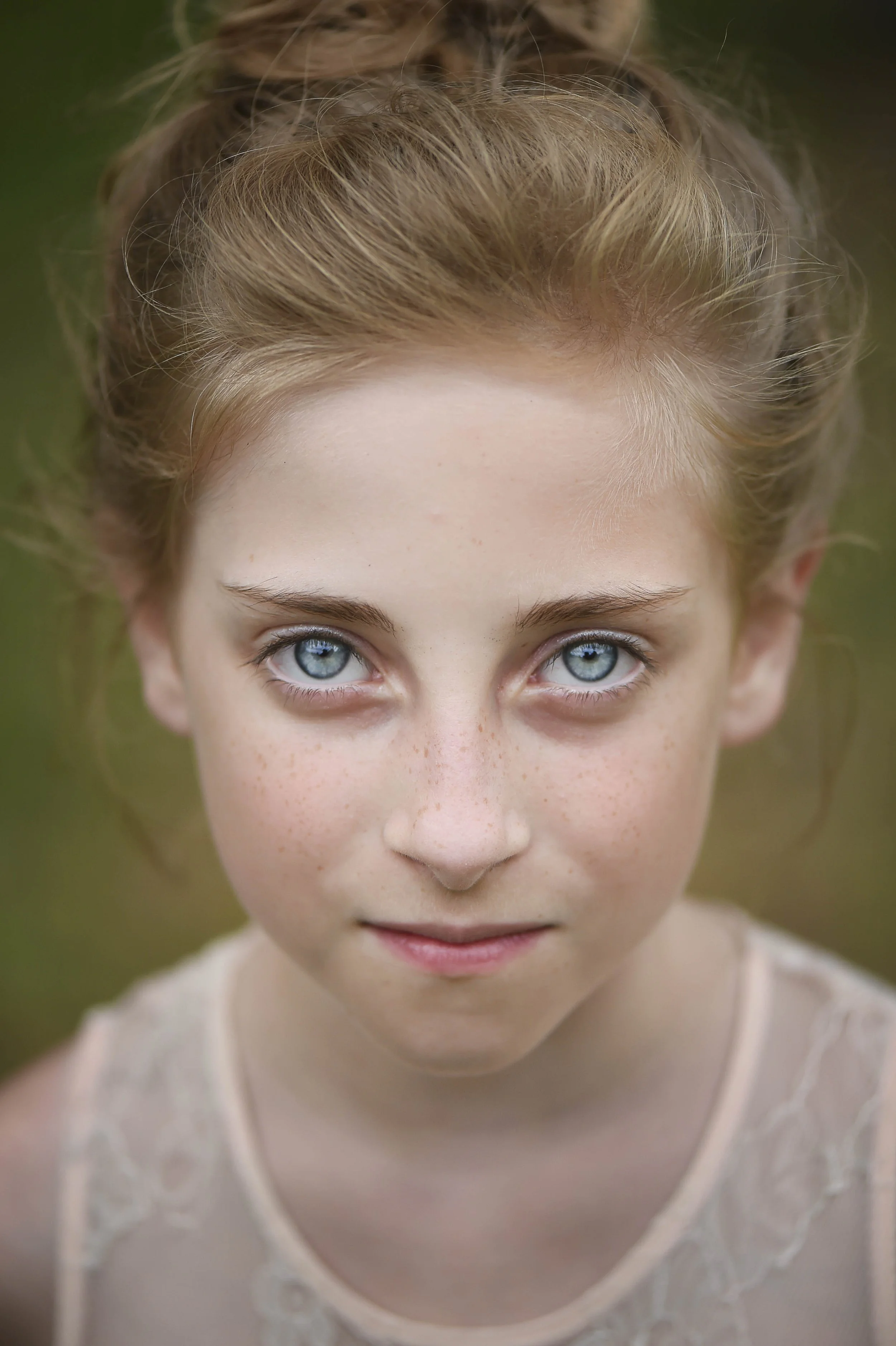 Close-up of a girl with blue eyes and ginger hair tied up in a bun, wearing a light-colored lace top, outdoors with a blurred green background.