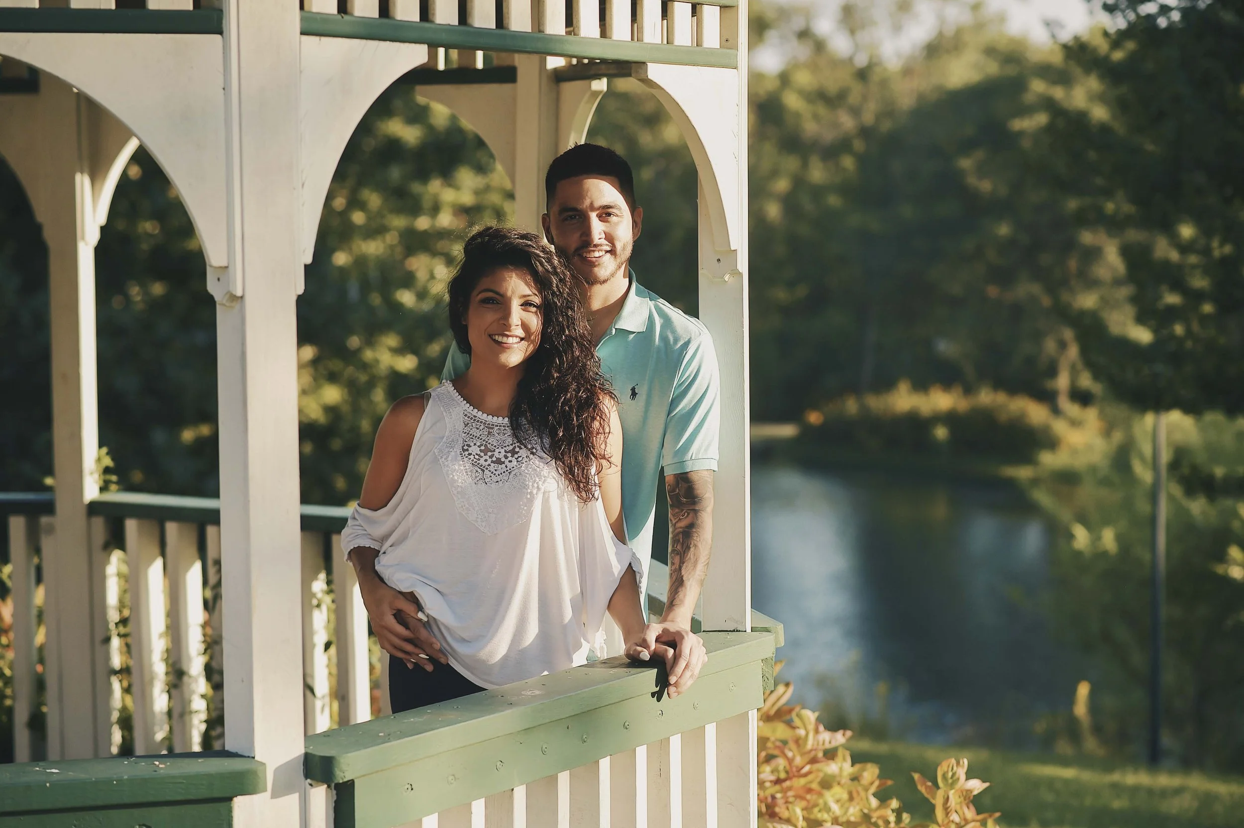 Two smiling people, a woman with curly dark hair wearing a white lace top and a man with short dark hair and a tattoo on his arm wearing a light blue polo, standing on a green and cream-colored gazebo with a lake and trees in the background during da