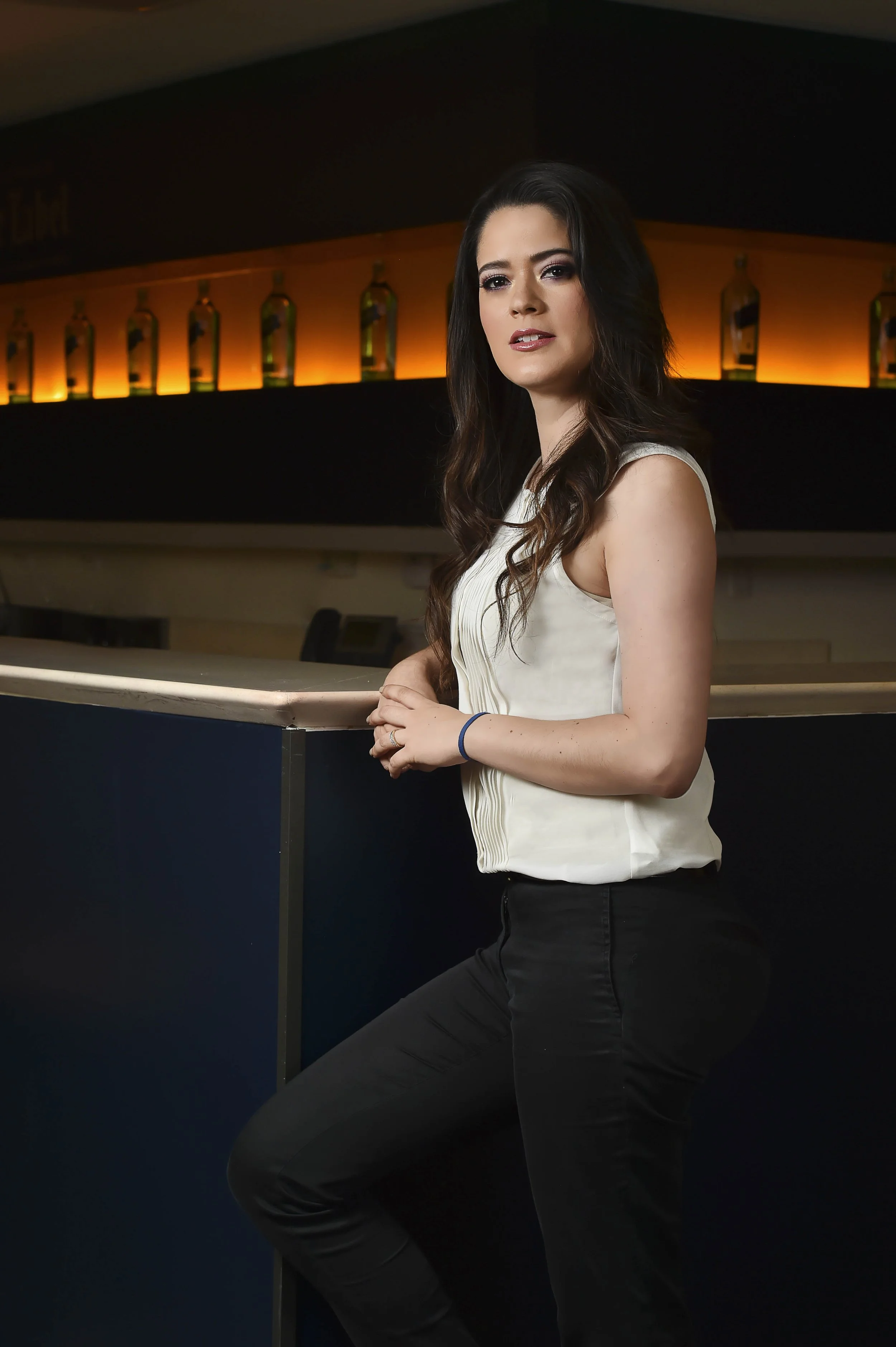 A woman with long dark hair, wearing a sleeveless cream top and black pants, leaning against a bar counter in a dimly lit bar or lounge.