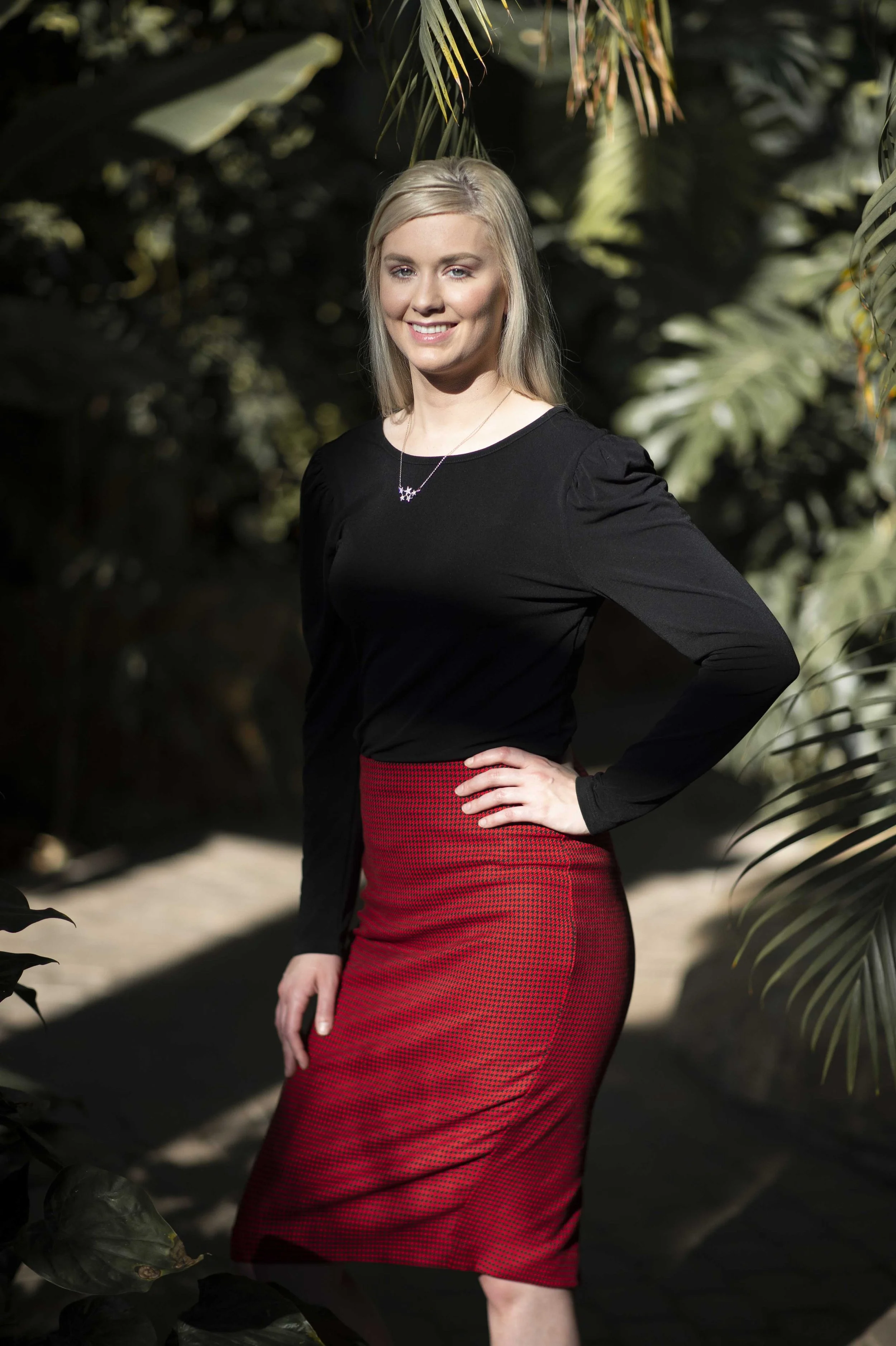 A woman with blonde hair and fair skin stands outdoors in a sunny setting, wearing a black long-sleeve top and a red, fitted skirt with a textured pattern. She is smiling and has her right hand on her hip, with lush green foliage in the background.