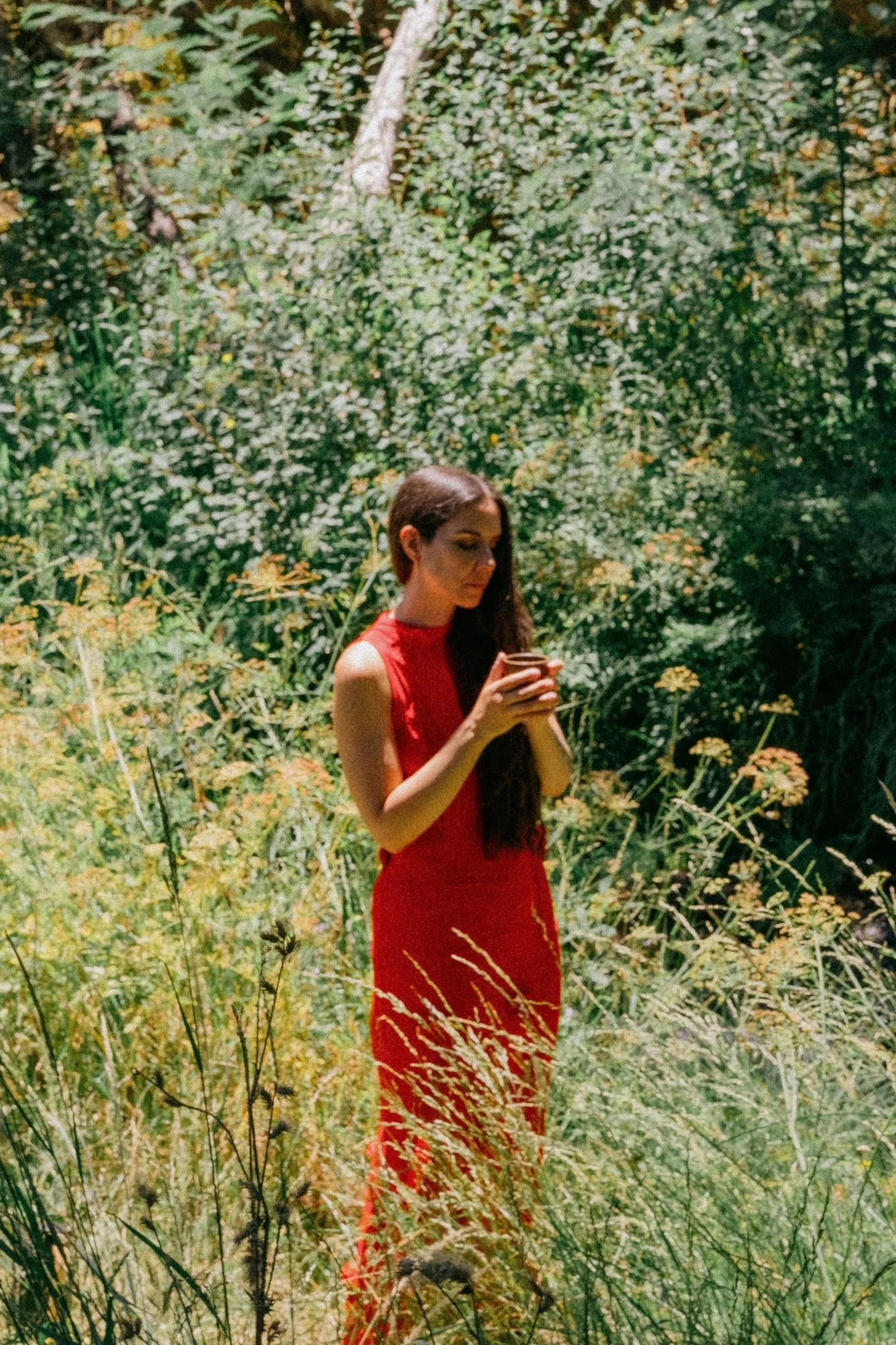 A woman with long dark hair wearing a red dress is standing outdoors in a lush green garden, holding a glass of drink in her left hand, surrounded by tall grass and plants.