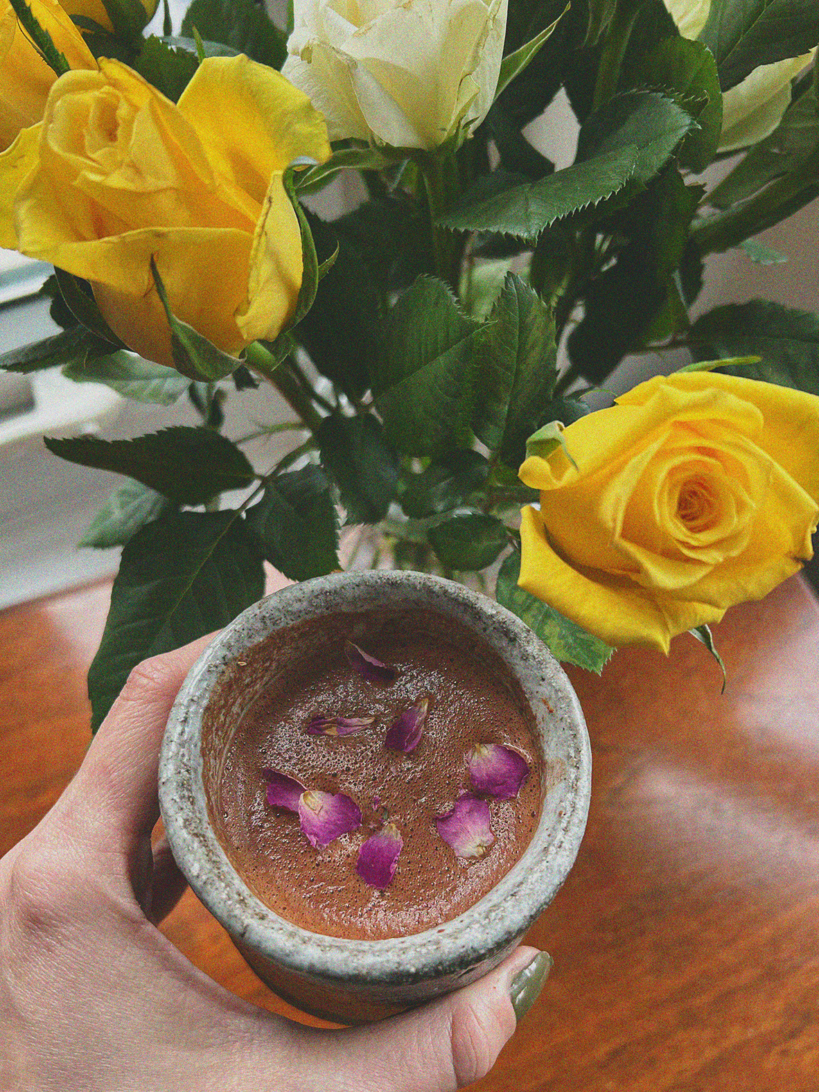 A hand holding a small stone cup filled with a beverage, garnished with pink flower petals, with a bouquet of yellow and white roses in the background.