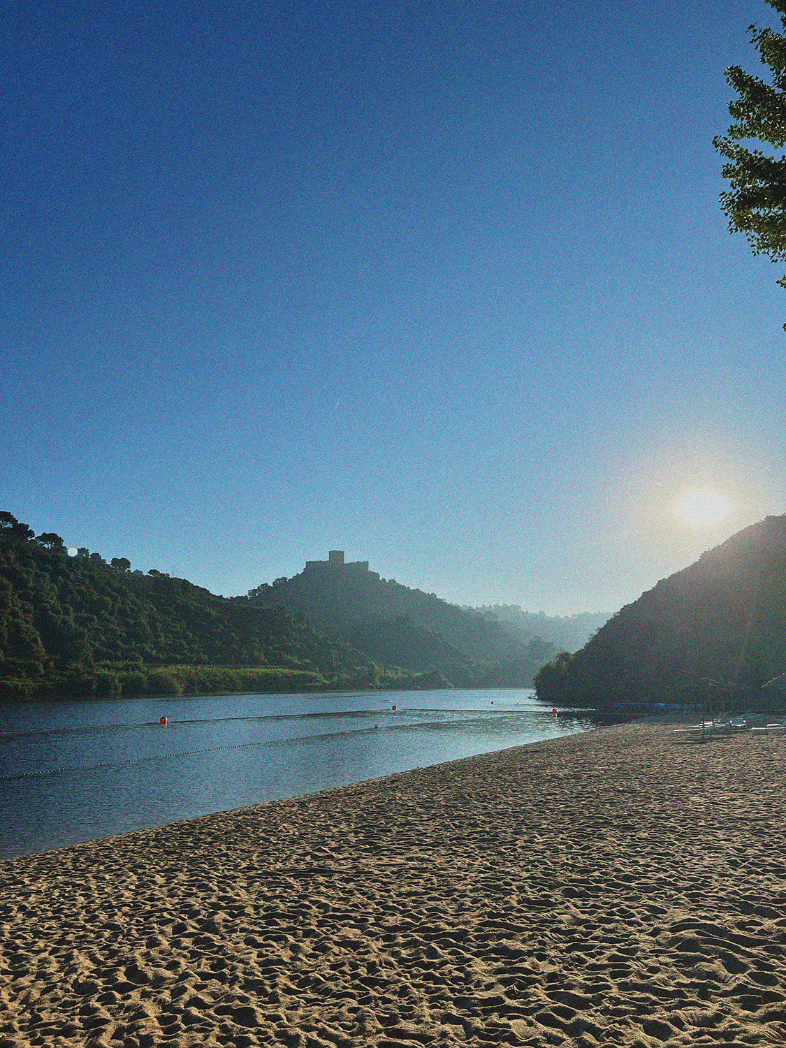 Sunrise over a calm river surrounded by green hills and a sandy beach in the foreground.
