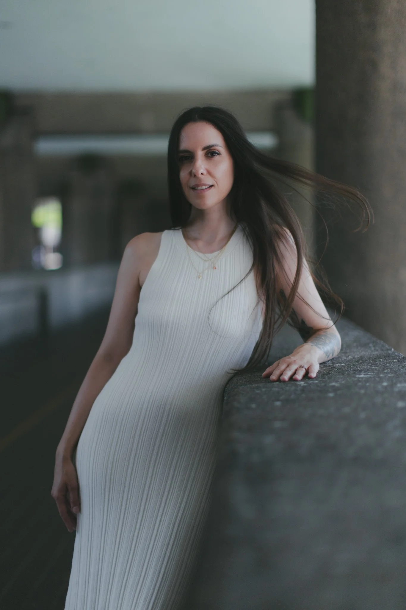 A woman with long dark hair wearing a sleeveless white pleated dress, standing outdoors, leaning against a concrete railing, with an urban background.