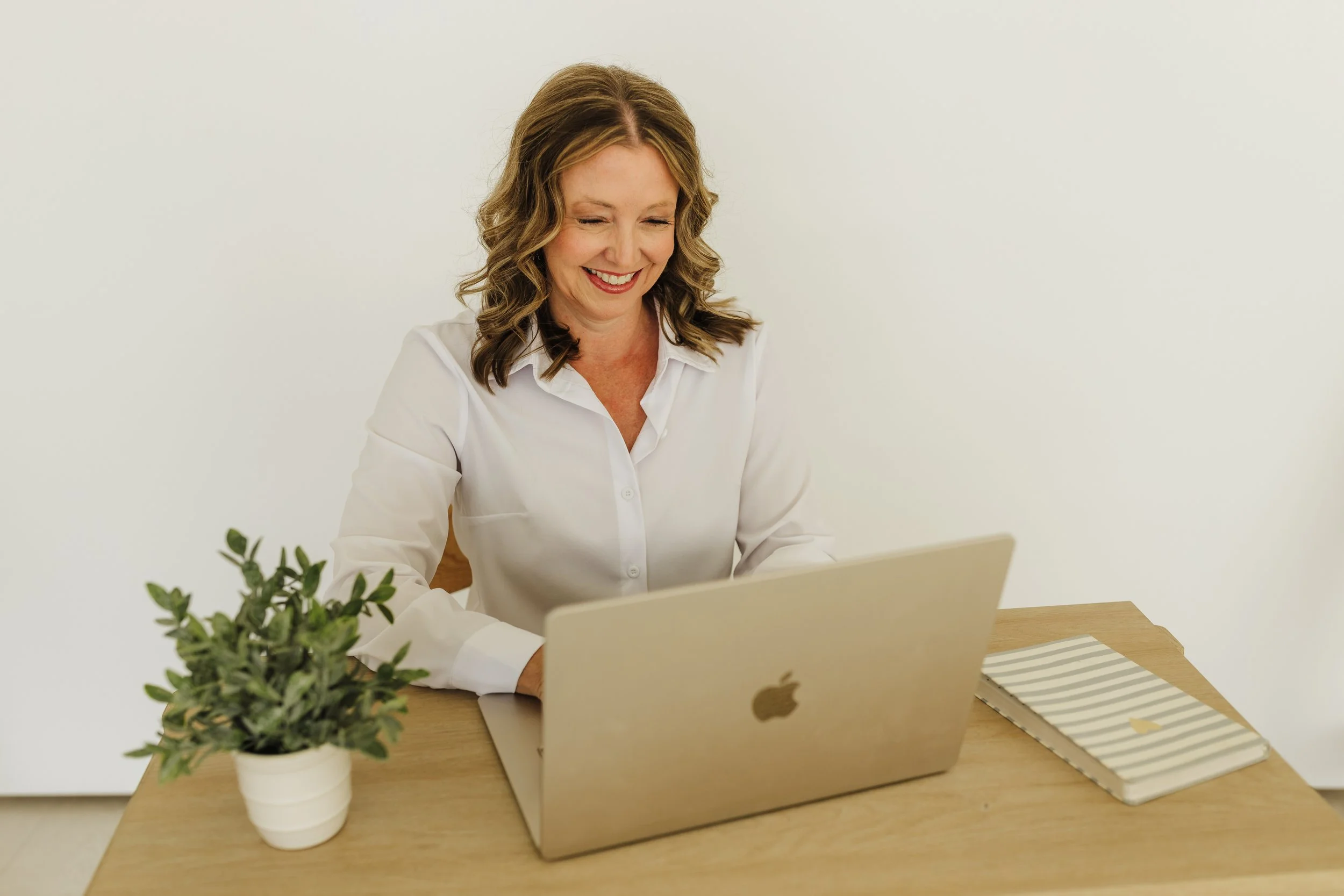 A woman with shoulder-length wavy hair, smiling while looking at a laptop on a wooden table, with a small potted plant and a notebook beside it, against a plain white background.