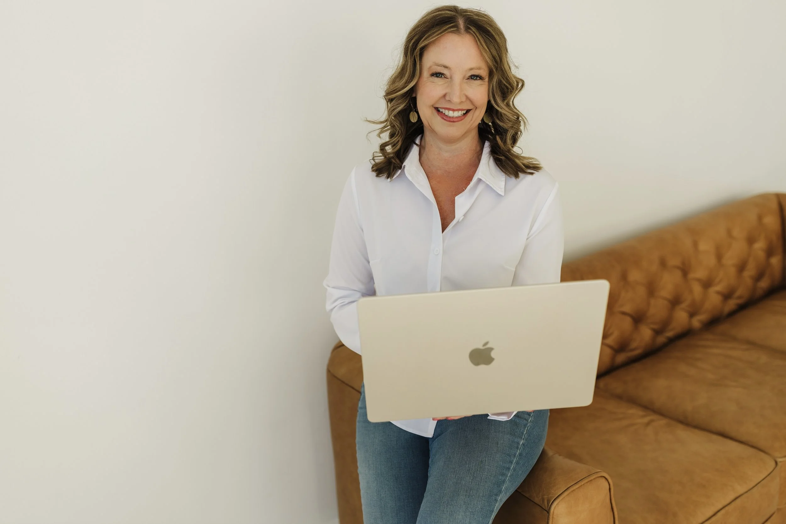 A woman with wavy light brown hair, dressed in a white button-up shirt and jeans, smiling while holding a silver MacBook laptop, sitting on a brown leather couch against a plain white wall.