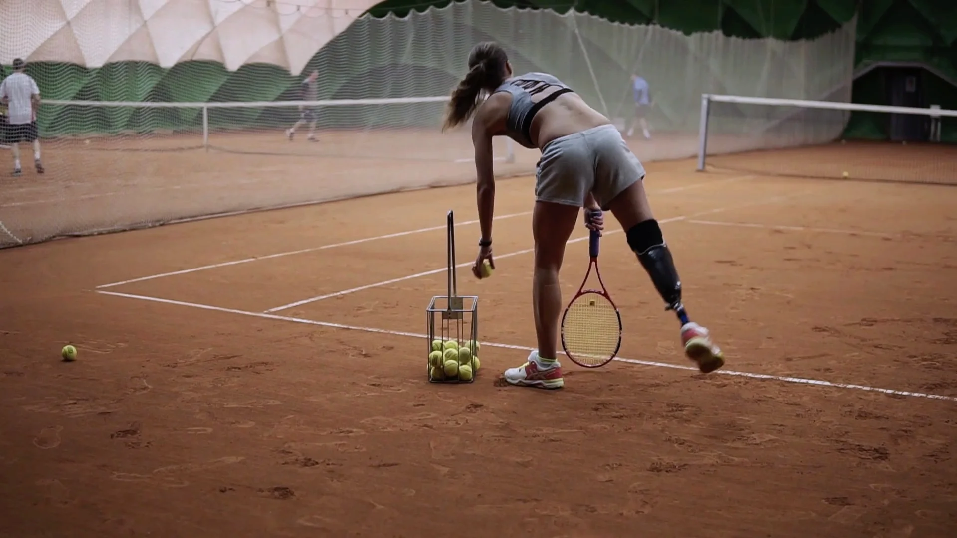 videoblocks-two-beautiful-girls-practicing-the-handicapped-girl-in-short-shorts-takes-the-ball-from-the-basket-and-makes-the-first-serve-of-the-ball-indoor-tennis-court-back-view_rhxke7nkpm_thumbnail-full01.jpg
