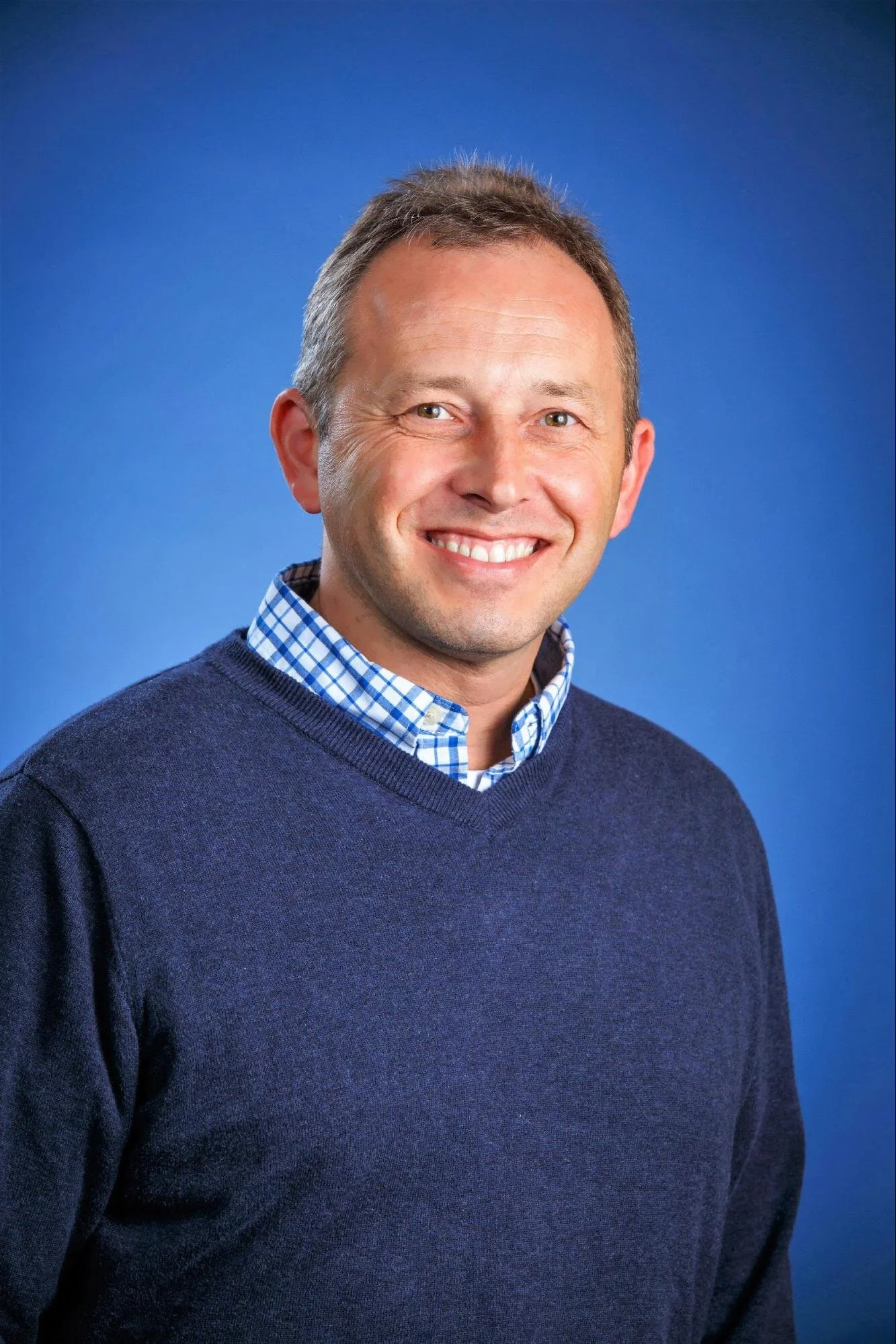A smiling man with short brown hair wearing a blue sweater and a blue checkered collared shirt against a blue background.