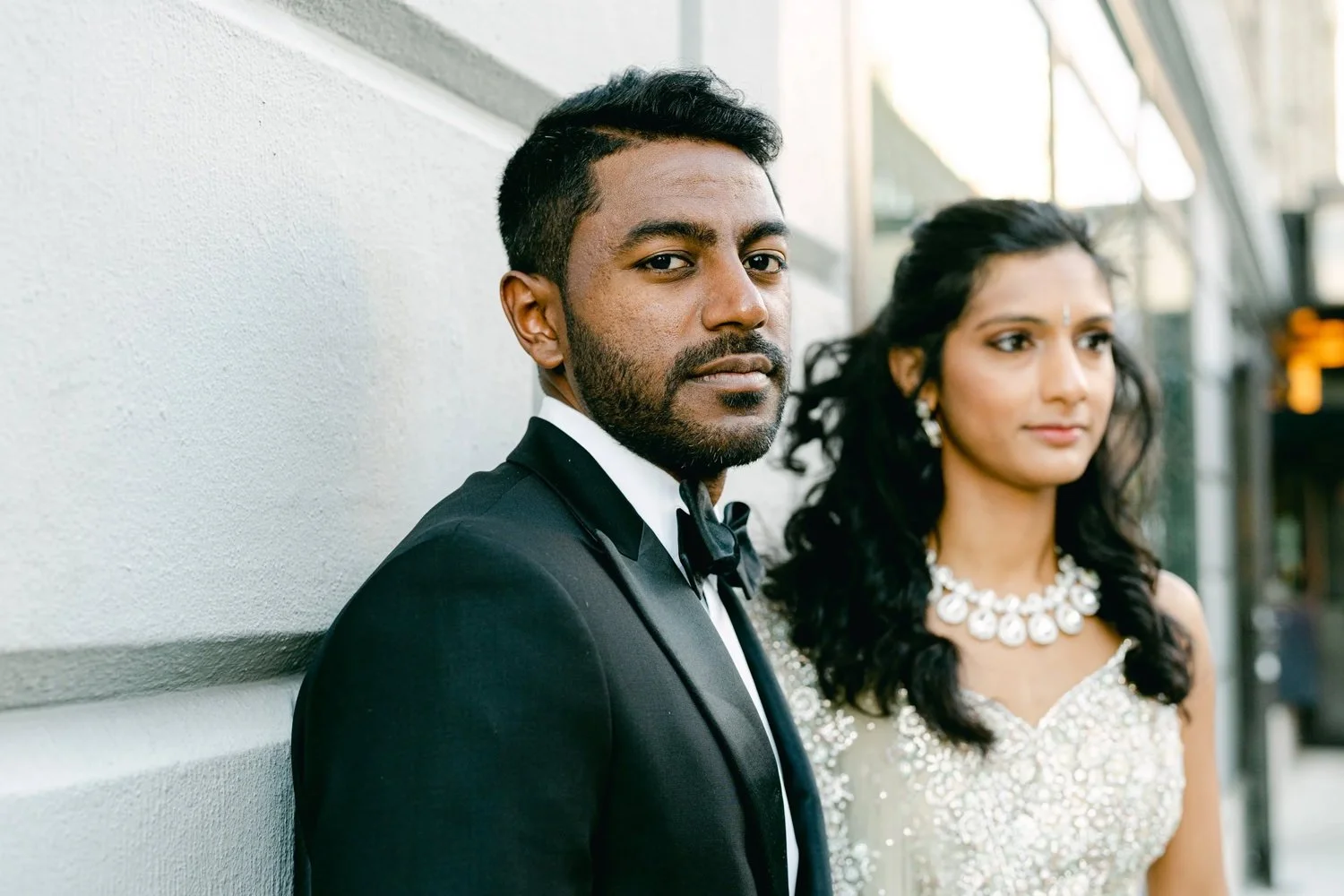 A man in a black tuxedo standing in front of a woman in an elegant dress, both looking slightly away from the camera, with an urban background.
