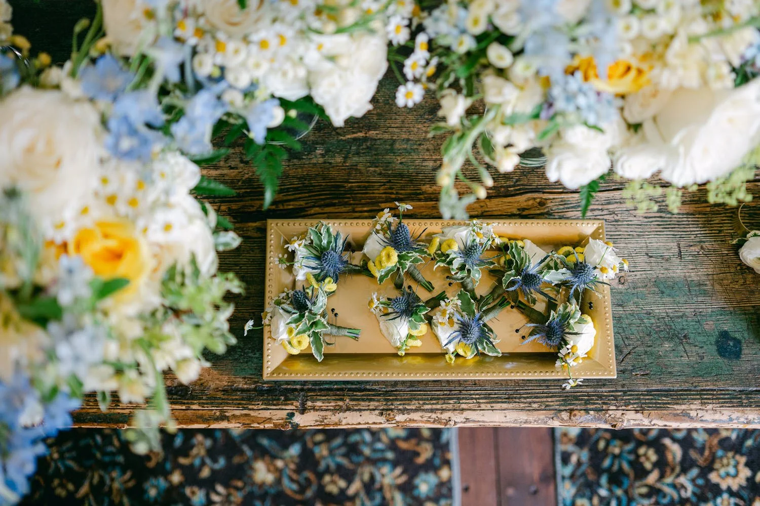 A top-down view of a rectangular tray with floral boutonnieres on a wooden table, surrounded by bouquets of flowers.