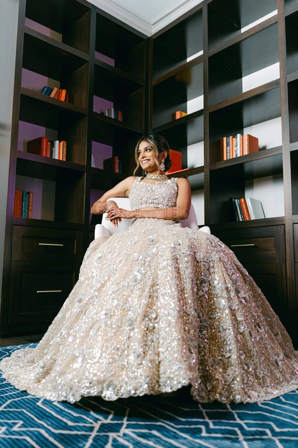 A woman in an ornate, sparkling gown sits confidently on a white chair in front of a dark wooden bookshelf filled with colorful books, smiling and looking to her left.