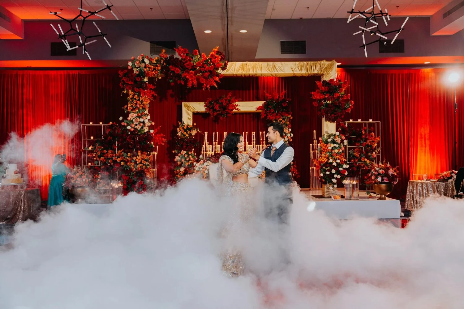 A bride and groom dance together at their wedding reception, surrounded by red and pink flowers, candles, and red curtains, with a cloud-like fog on the floor.
