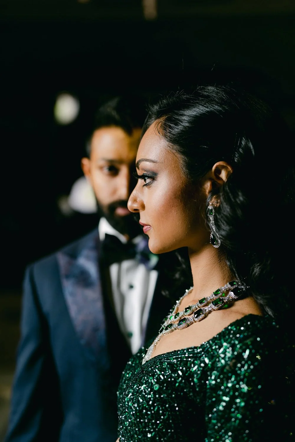 Elegant woman in a green, sequined gown with jewelry standing in front of a man in a tuxedo, both looking serious.