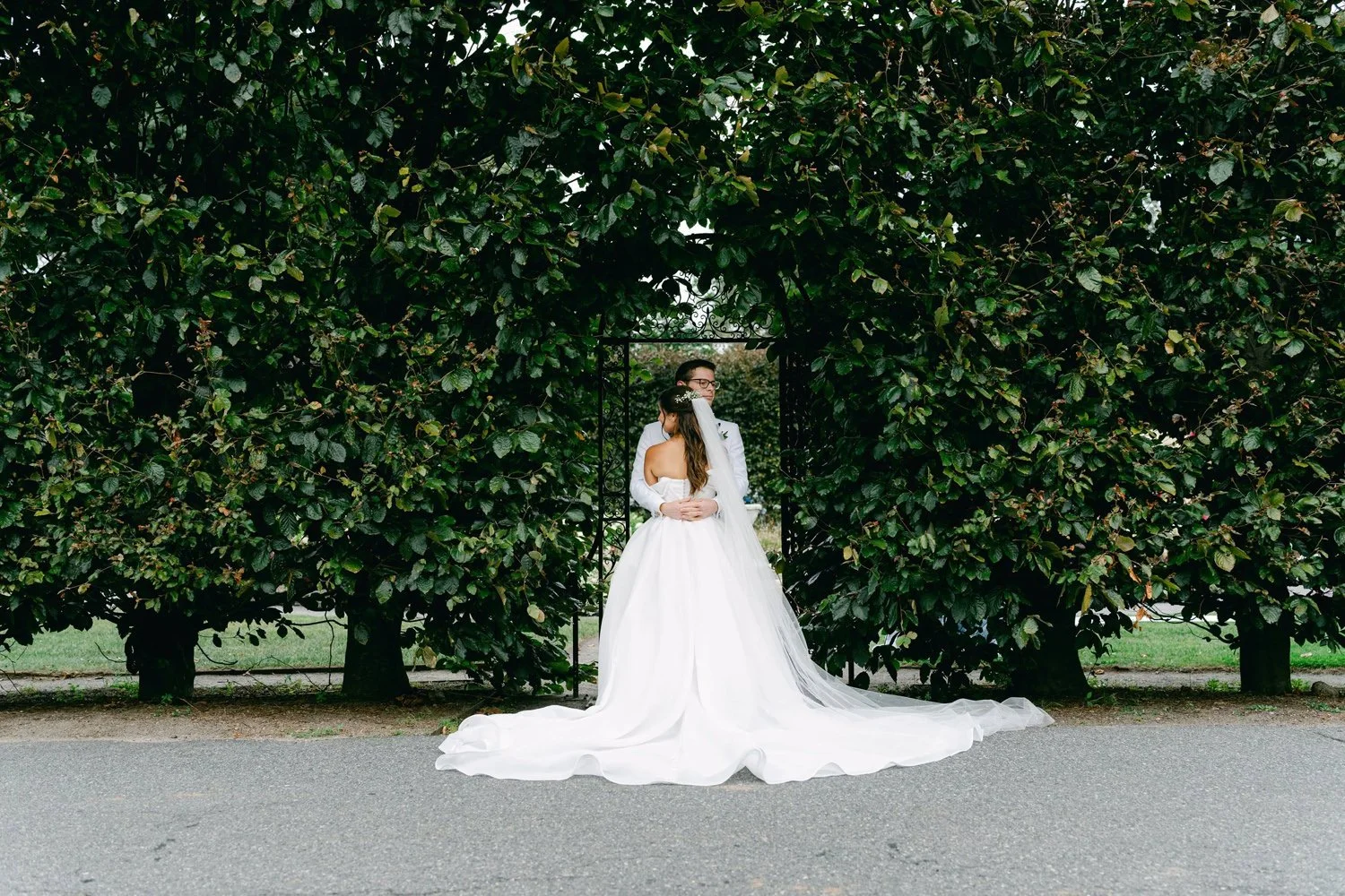A bride and groom embracing in front of a green hedge, with a decorative black gate behind them. The bride is wearing a white wedding gown and veil, and the groom is in a white suit with glasses.