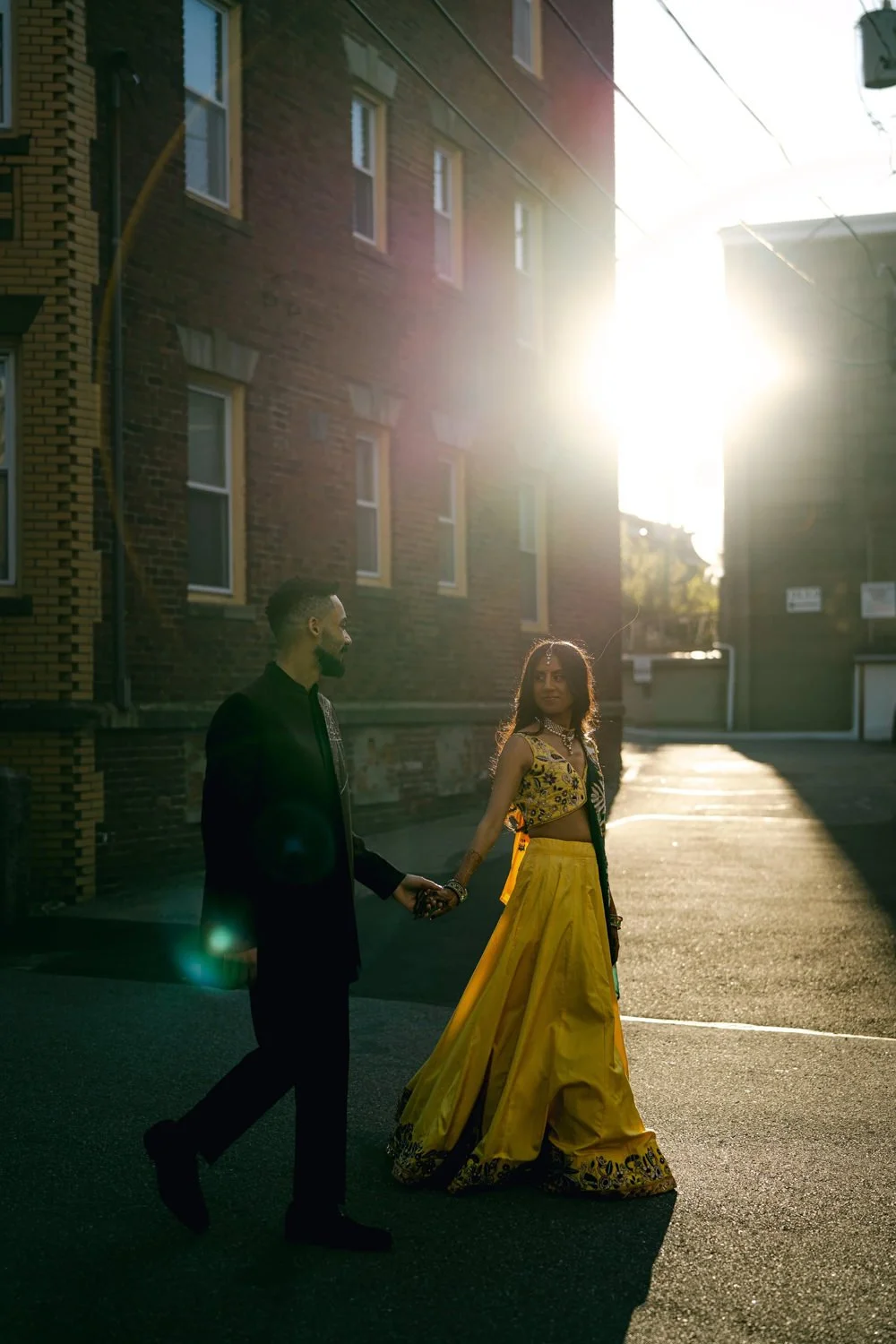 A man and a woman dressed in traditional Indian attire are walking hand-in-hand in an alleyway during sunset, with the sunlight creating a dramatic glow behind them.