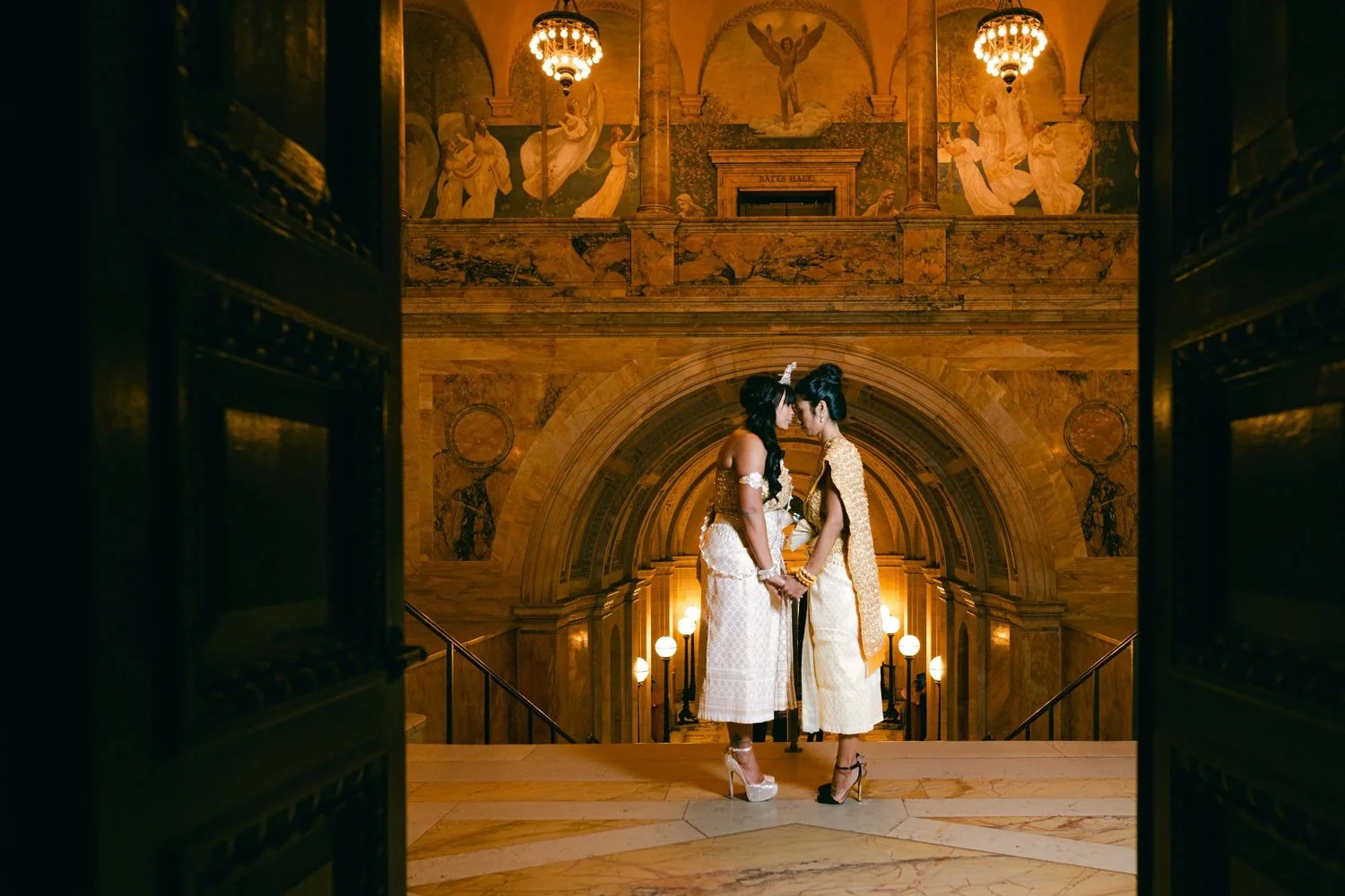 Two women dressed in traditional Thai attire standing close together in an elegant, grand hall with ornate architecture and golden decor, holding hands and touching foreheads, surrounded by warm lighting and historic murals.