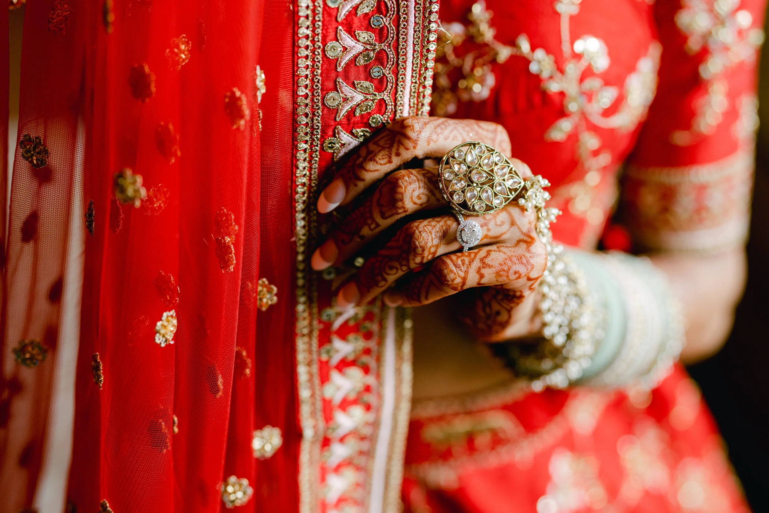 Close-up of a woman's hand with intricate henna and large jewelry rings, resting on red embroidered traditional attire.