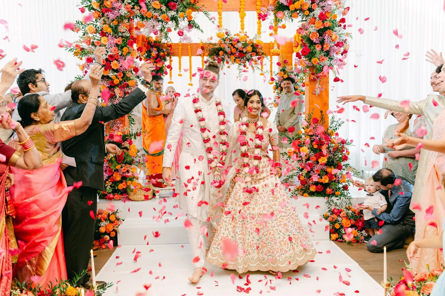 A wedding celebration with a bride and groom walking hand in hand under a floral arch, surrounded by guests throwing pink flower petals.