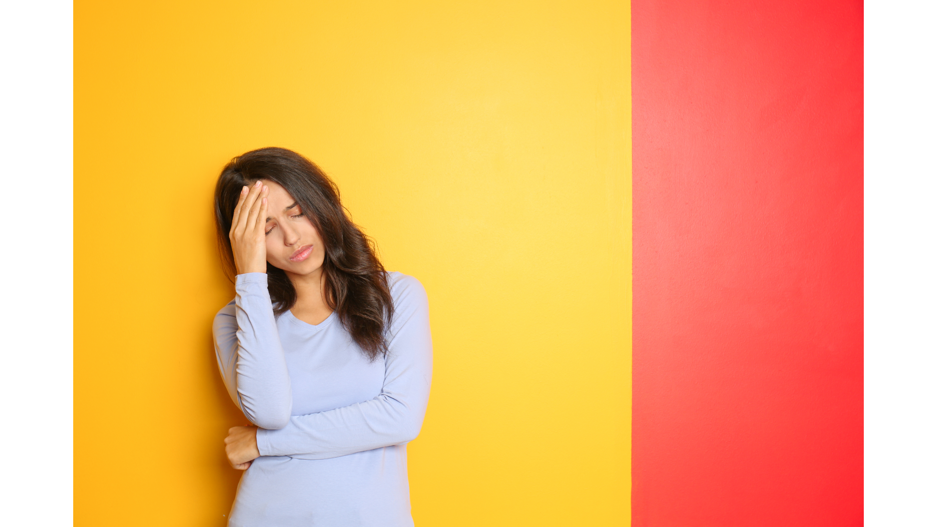 A woman standing and leaning on a yellow wall. He head is in her hands and she looks stressed.