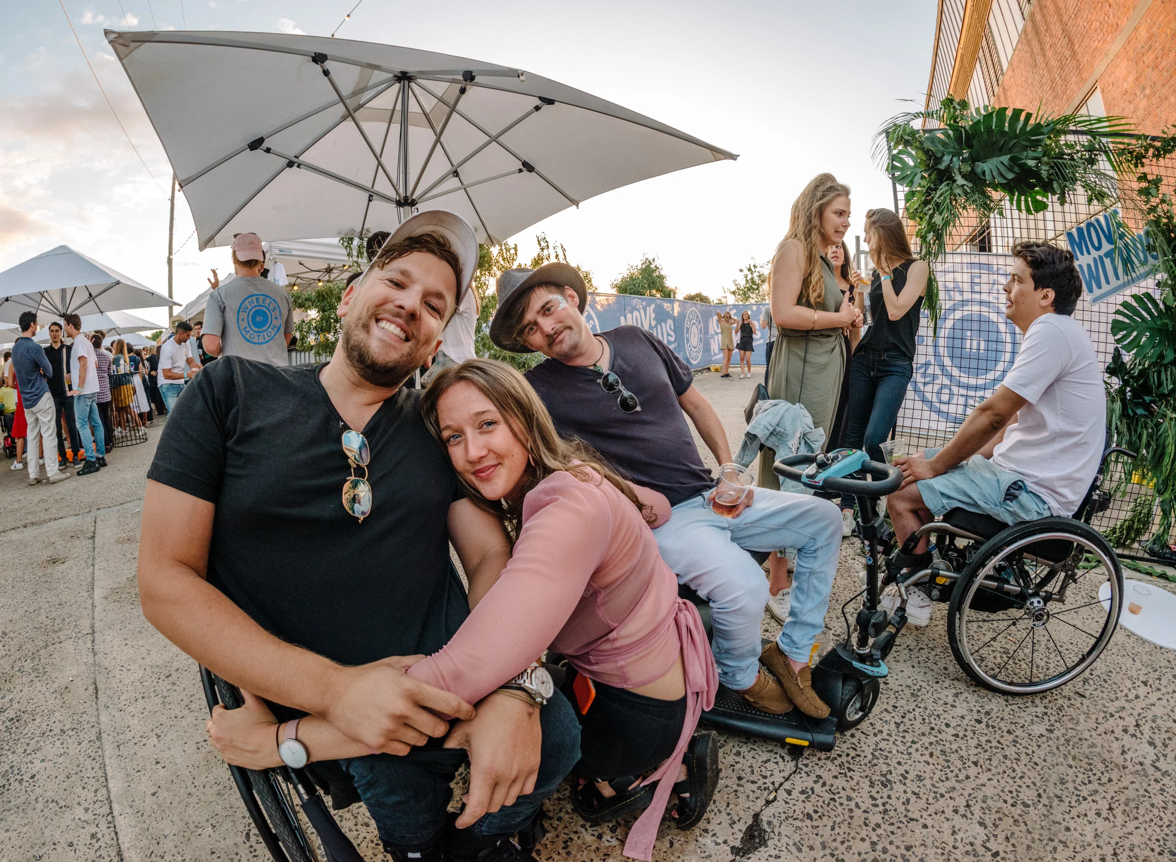 Image of 3 friends hugging. One uses a wheelchair, one uses a mobility scooter and one is squatting. There is another wheelchair user in the background. Everyone looks cool and happy!