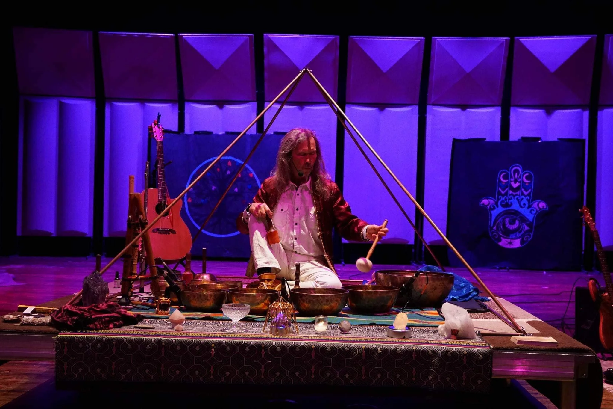 A person with long hair playing percussion instruments on stage, surrounded by musical instruments and decorations, with purple lighting and a backdrop featuring a blue emblem.