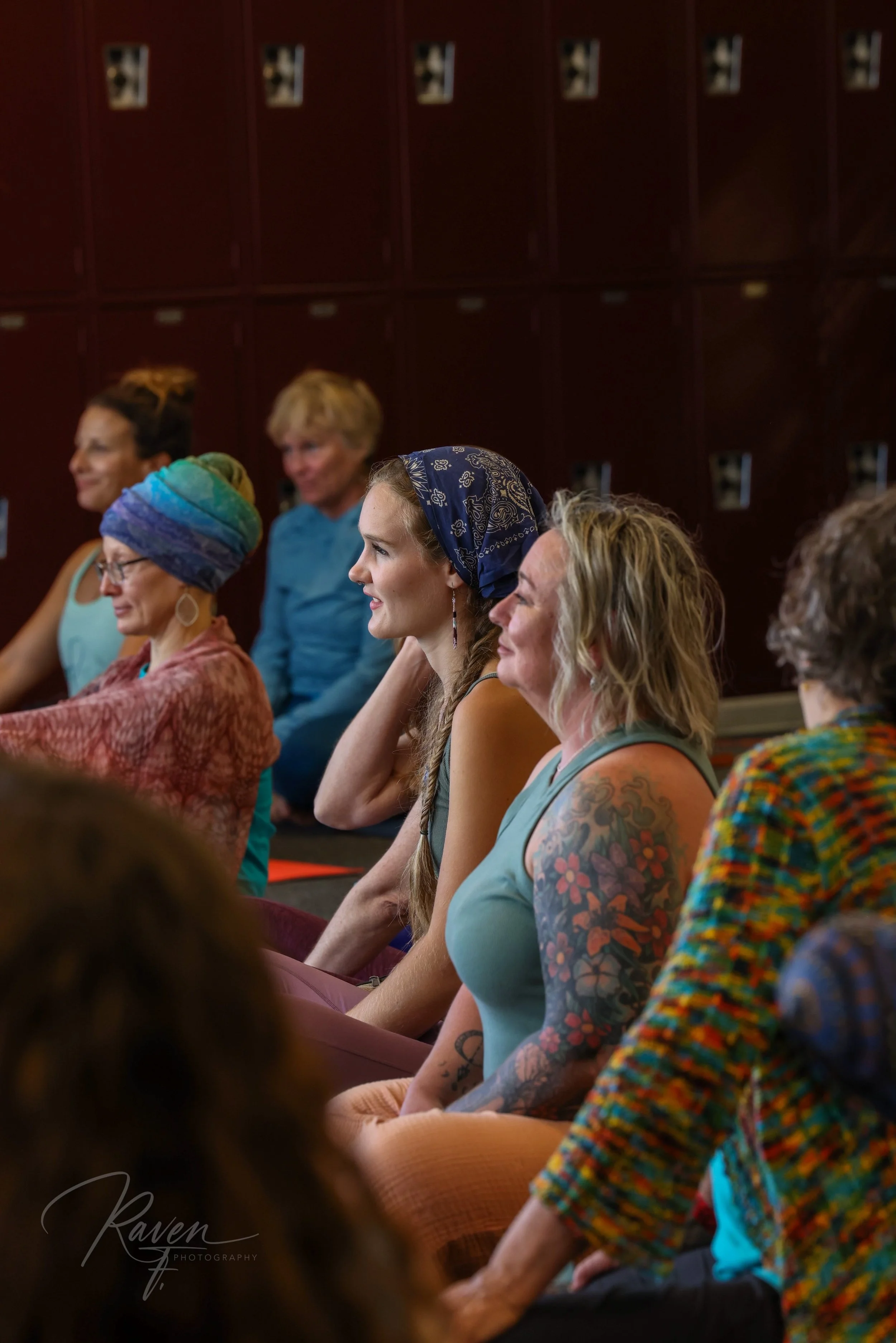 People sitting cross-legged in a yoga or meditation class in a room. They appear calm and focused, some with open eyes, others possibly meditating.
