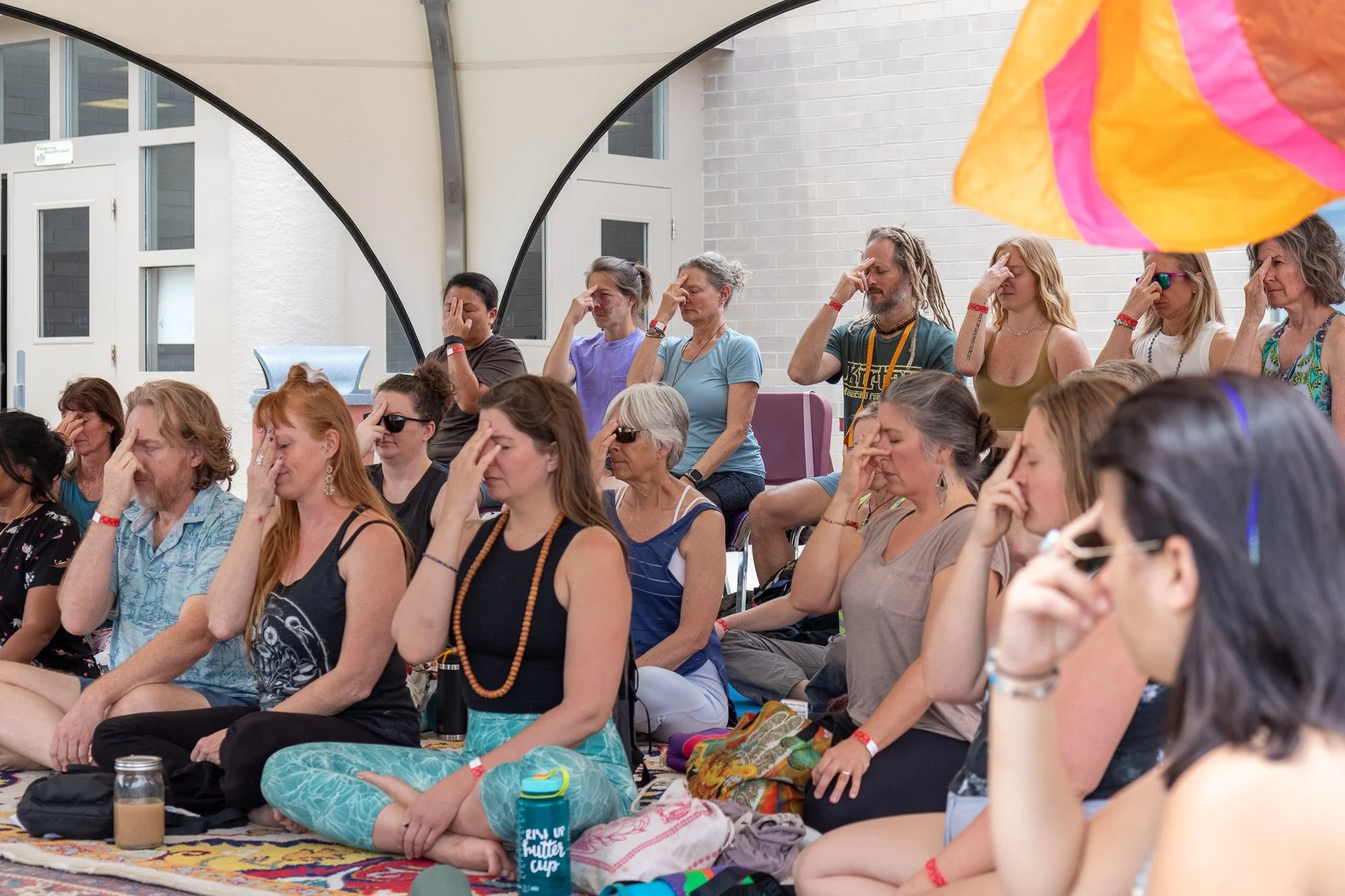 A group of people sitting cross-legged on the floor with their eyes closed and hands on their foreheads, participating in a meditation session inside a covered outdoor space.