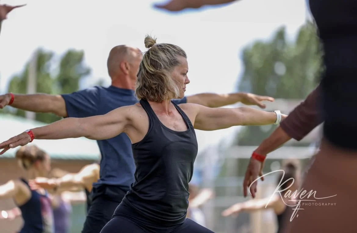 Group of people participating in outdoor yoga class, standing in Warrior pose with arms extended, women in black and men in blue shirts, trees and sky in background.