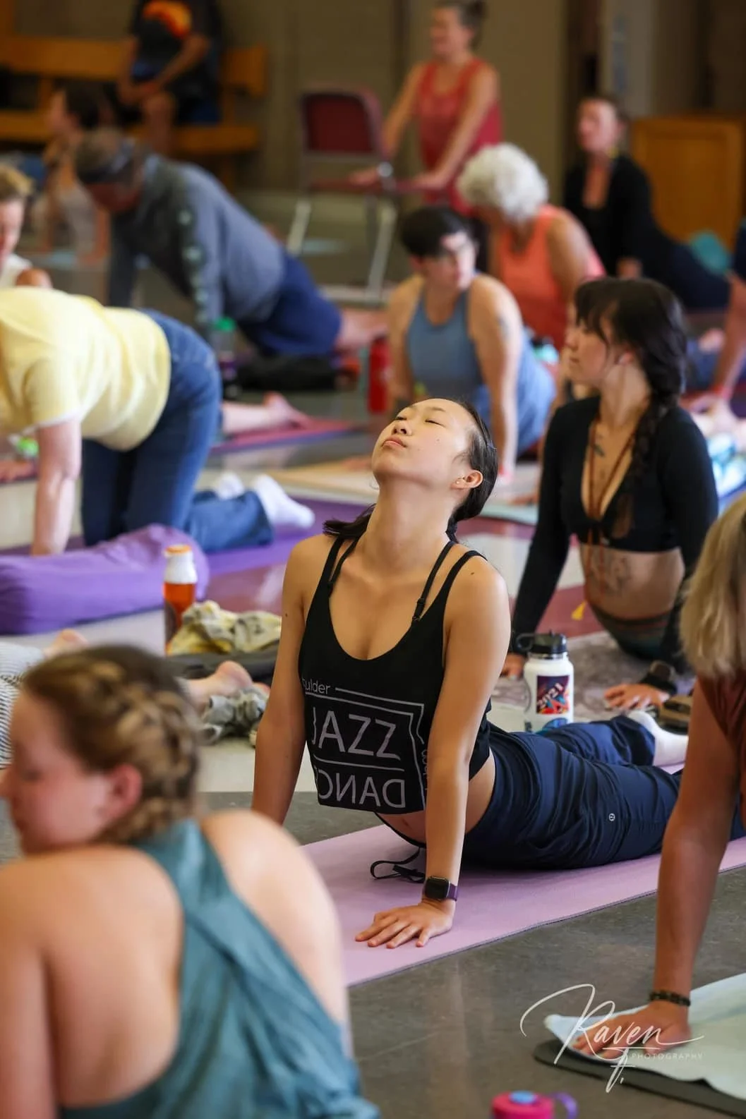 A group of people participating in a yoga or stretching class indoors, with a woman in a black tank top in foreground doing cobra pose, surrounded by others on yoga mats.