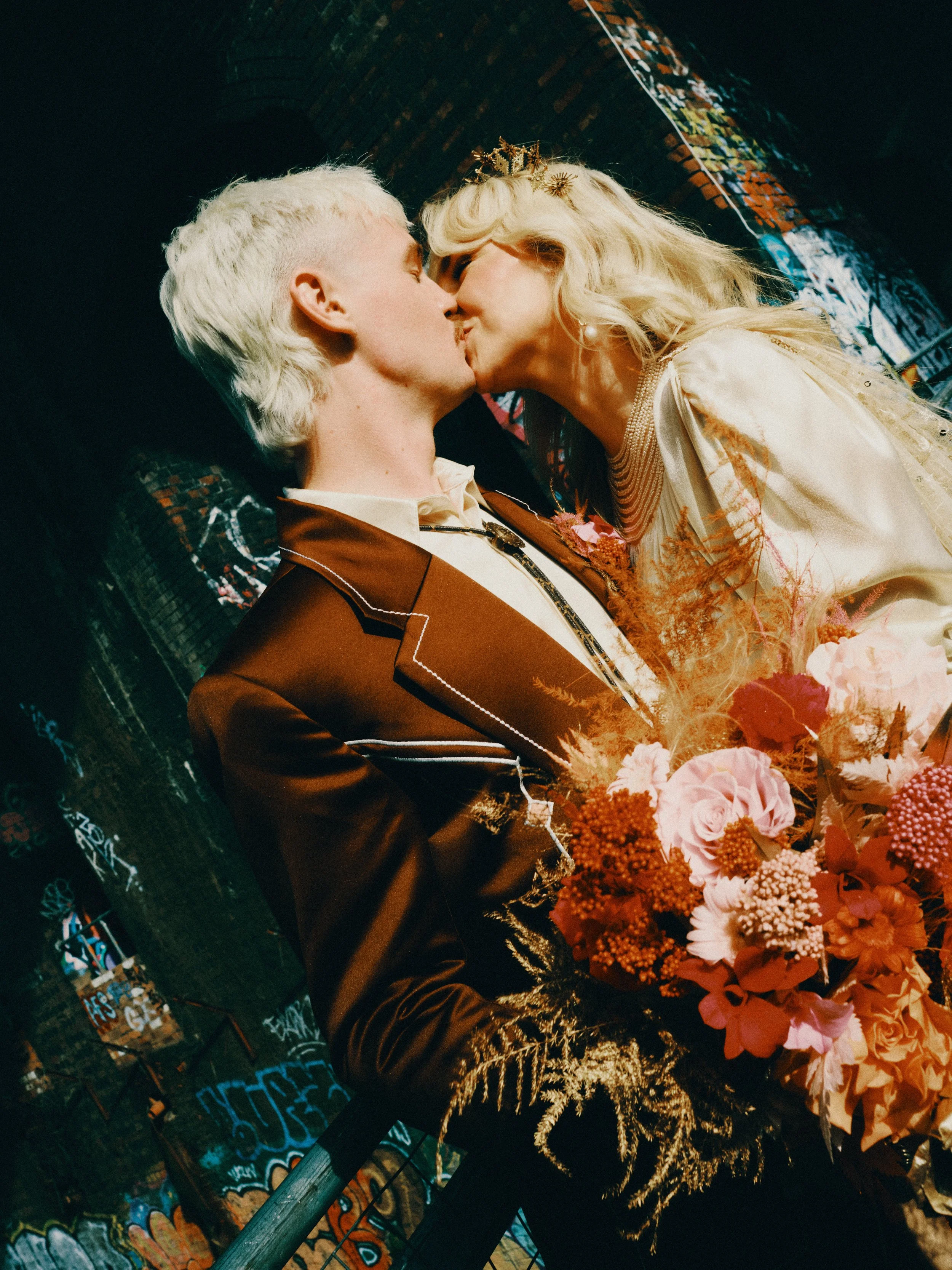 A man and woman sharing a kiss at a wedding, with the woman wearing a tiara and elegant dress, and the man in a suit, surrounded by a bouquet of pink and red flowers, with a graffiti-covered background. The Old Library, Digbeth.