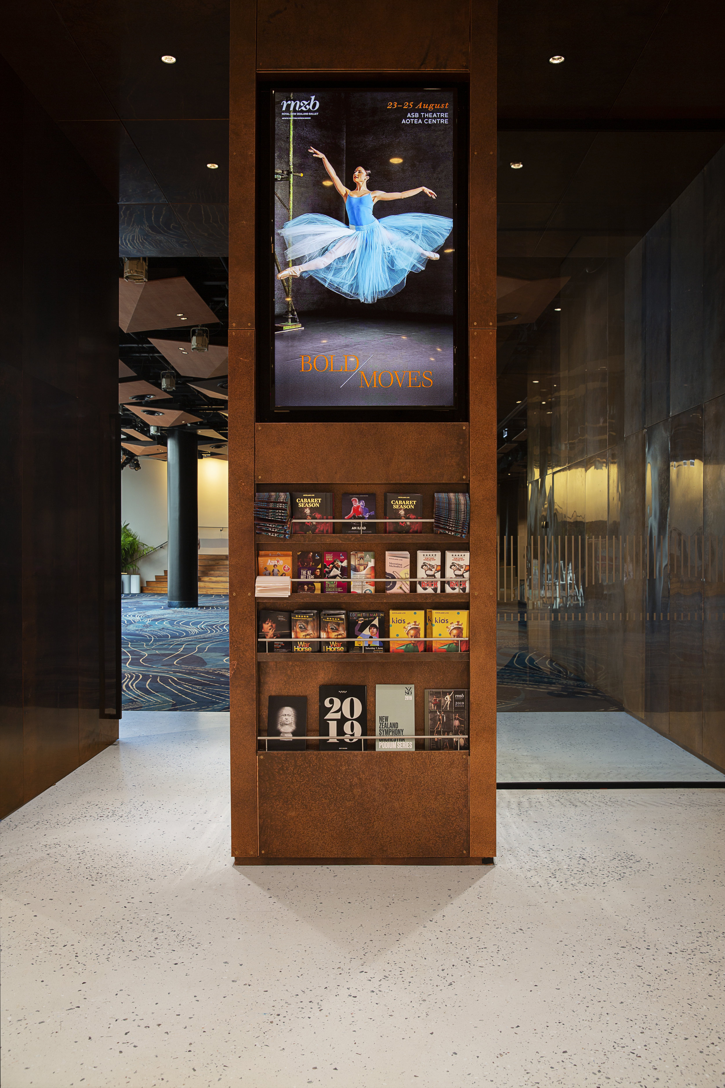 Interior view of a display stand featuring various booklets and programs, with a prominent digital poster above showing a ballet dancer in a blue tutu and the text 'Bold Moves', set in a modern building hallway with artistic flooring.