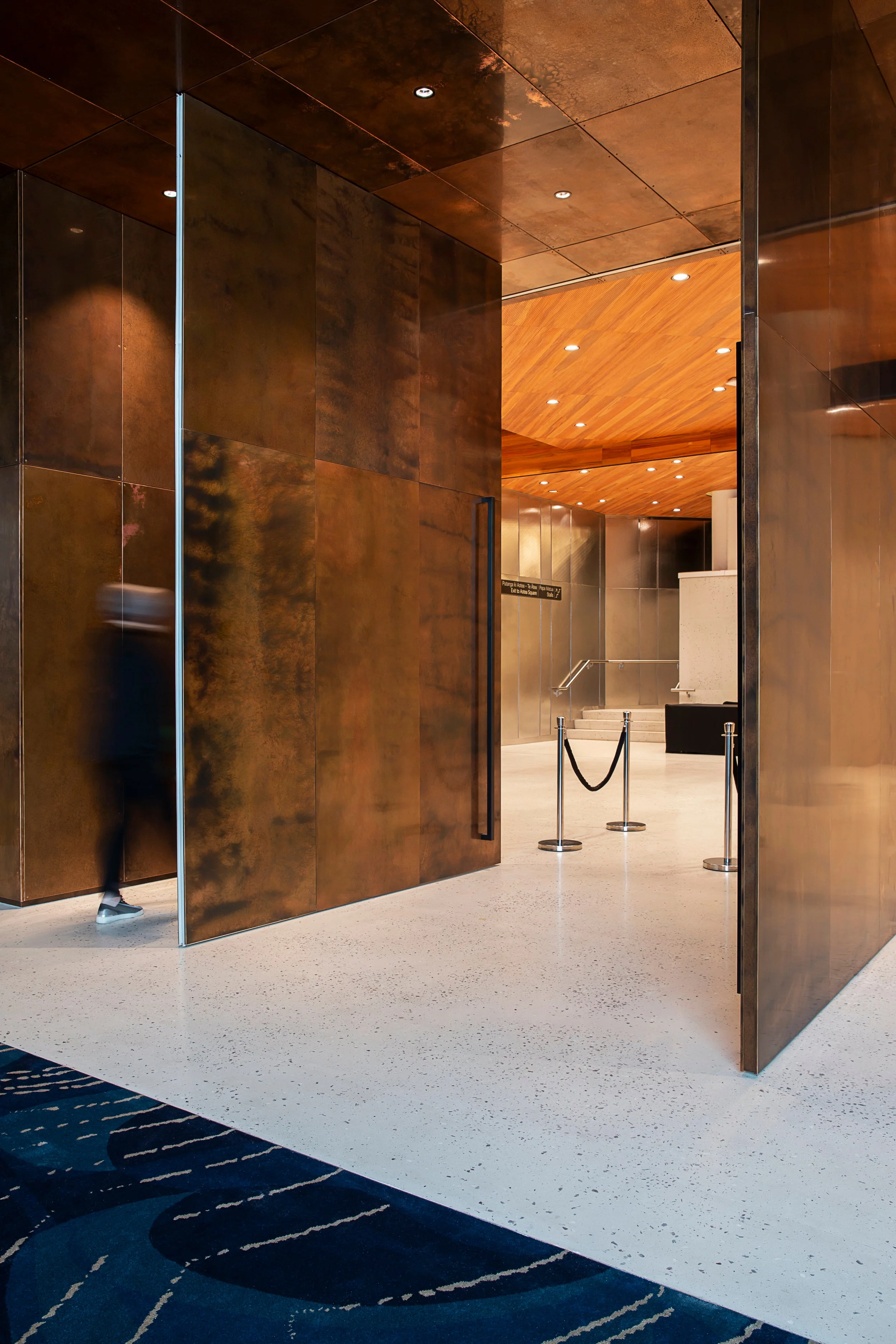 Modern interior with large copper doors, wooden ceiling, terrazzo flooring, and a blurred figure entering.