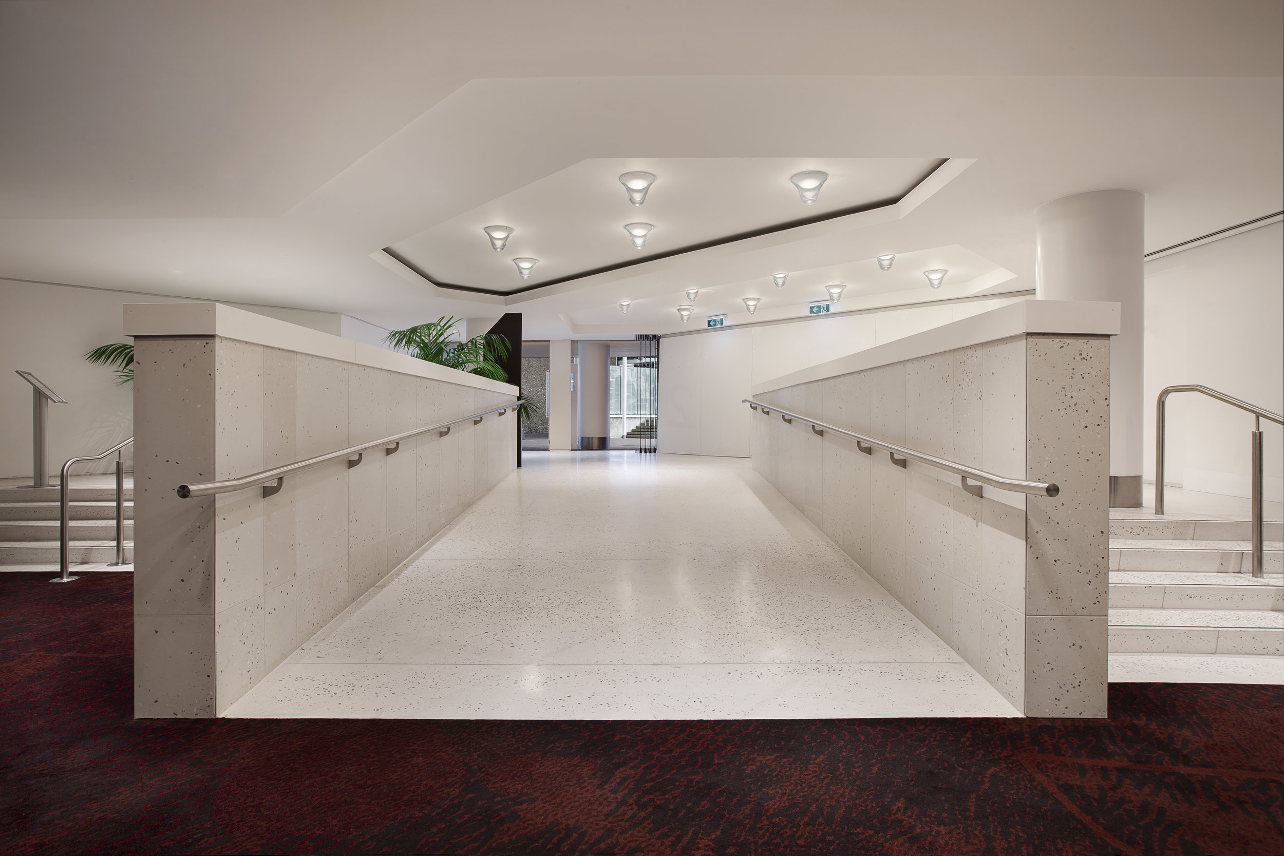 Modern indoor hallway with a white ramp and handrails, flanked by stairs, ceiling lights, and a carpeted floor.