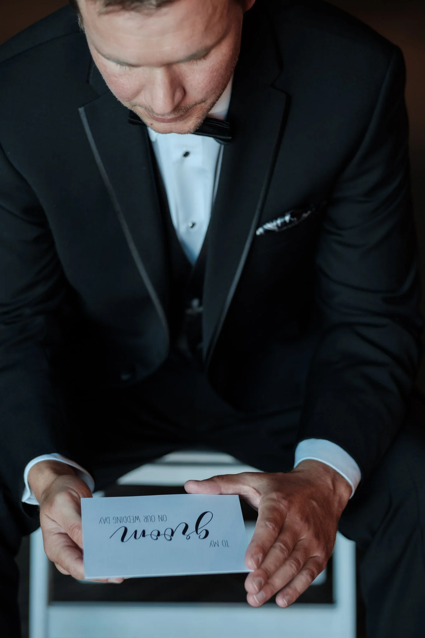 Groom reading letter from his bride before wedding begins with window light while dressed in his suit and bow tie.