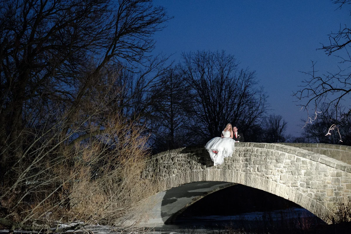 Night photo of bride sitting on the railing of a bridge snuggling with her husband.
