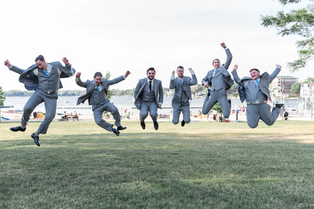 Groom and groomsmen jump for joy on the banks of lake oconomowoc in wisconsin