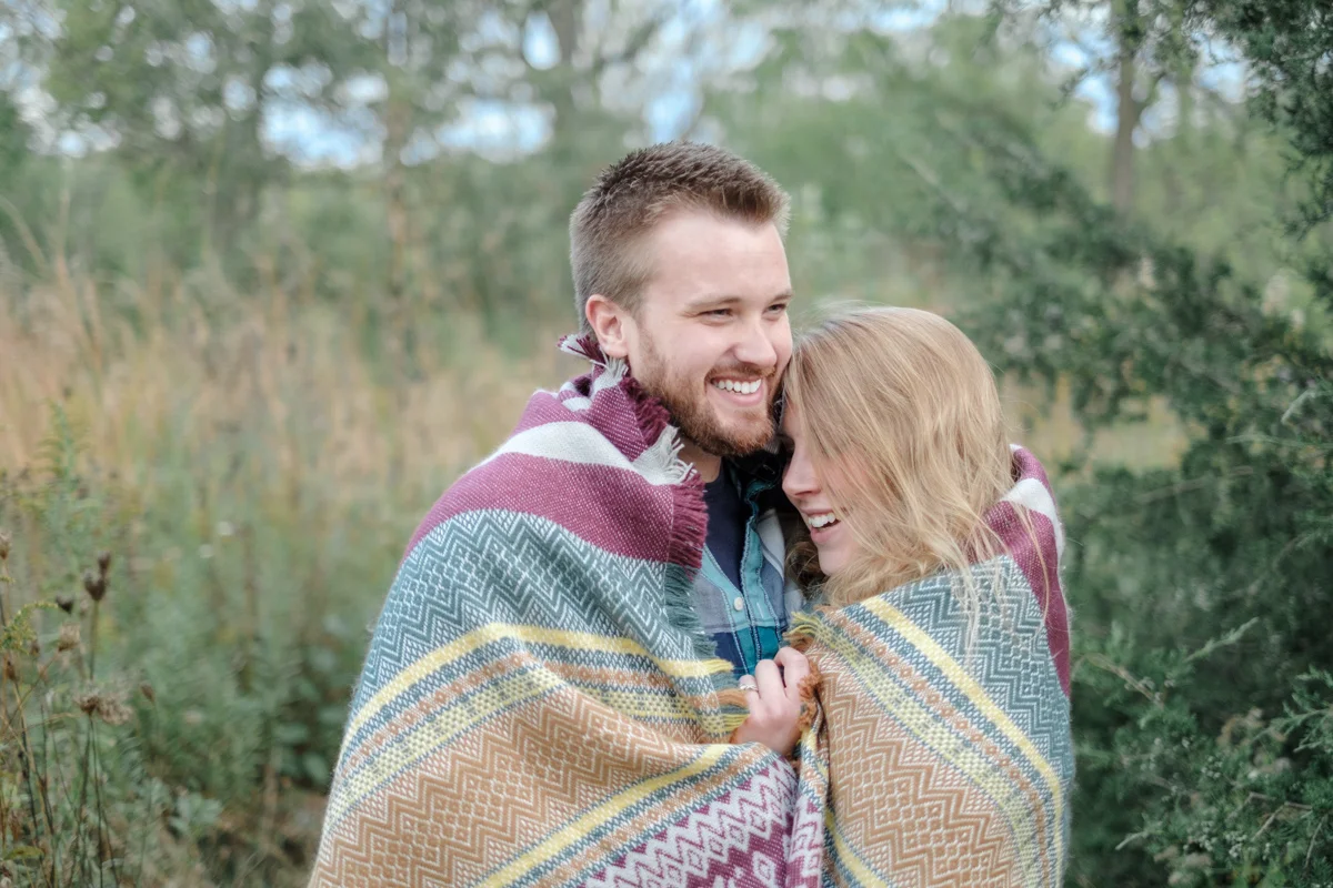 fall engagment portrait in macktown forest preserve with couple wrapped in blanket laughing