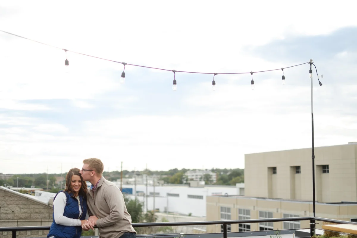 Engagement photo of Couple kissing under string lights at the rooftop bar of The standard on State in Rockford Illinois