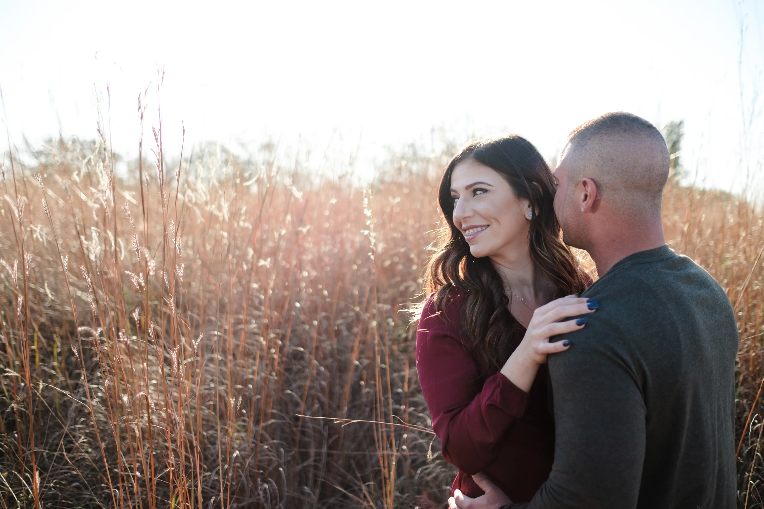 Bride with beautiful long dark hair looks away from groom with tall grass field behind them and the sun setting during golden hour in Rockford, Illinois.
