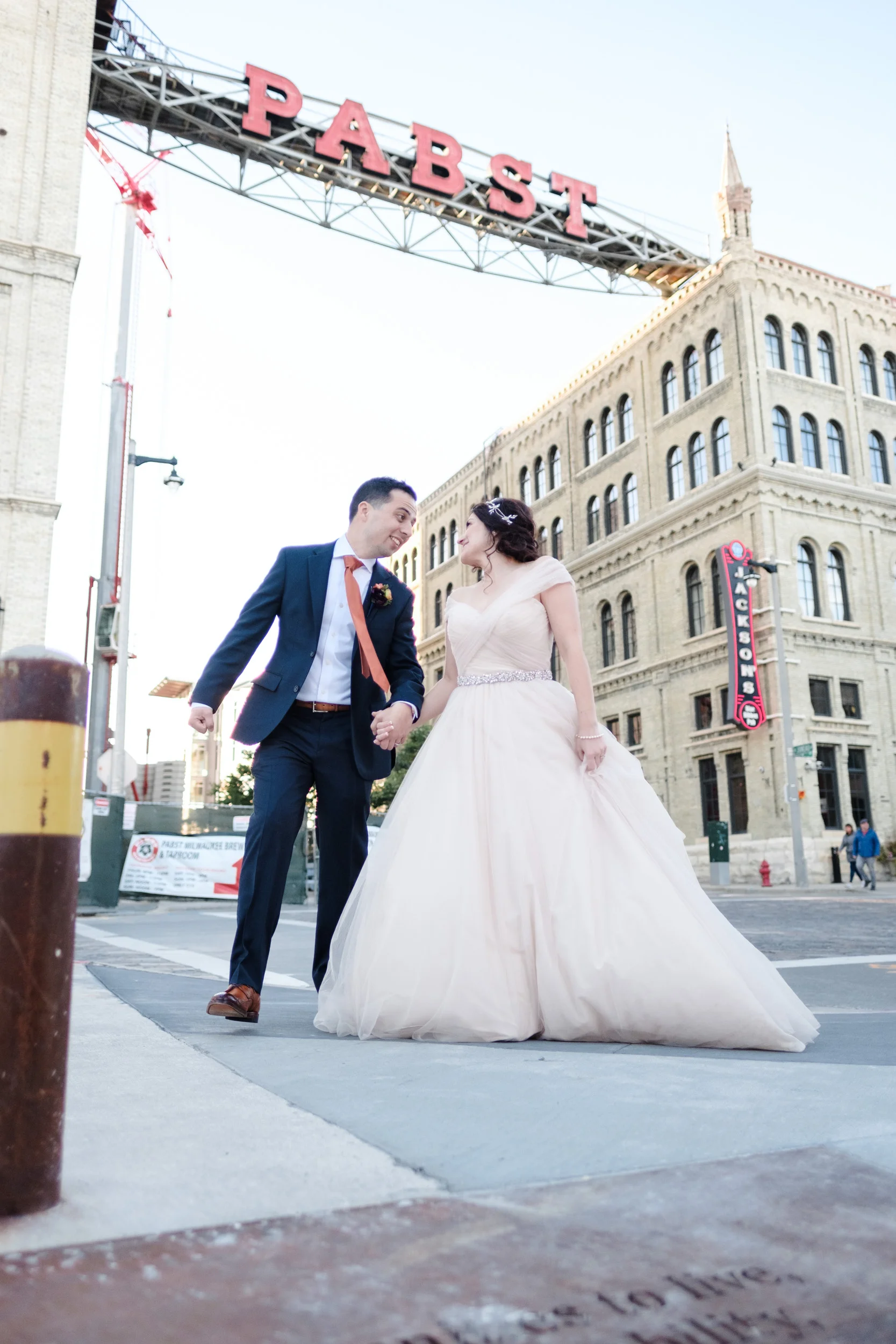 Bride and groom laugh and walk while holding hands in front of neon Pabst sign at Pabst best place in downtown Milwaukee Wisconsin.