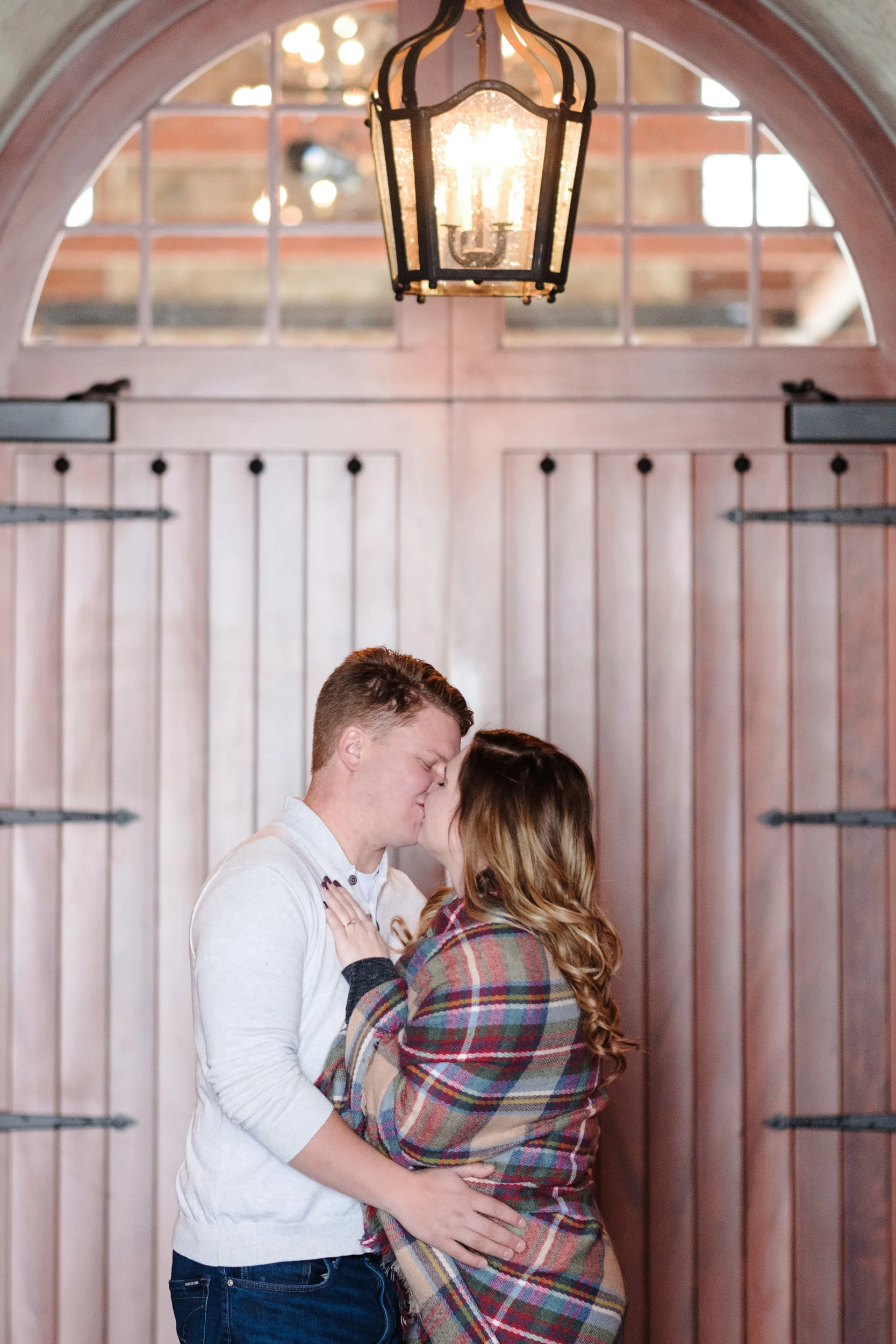 couple kisses with chandelier overhead at the pavilion at orchard ridge farms with large wooden doors behind them in brick tunnel.