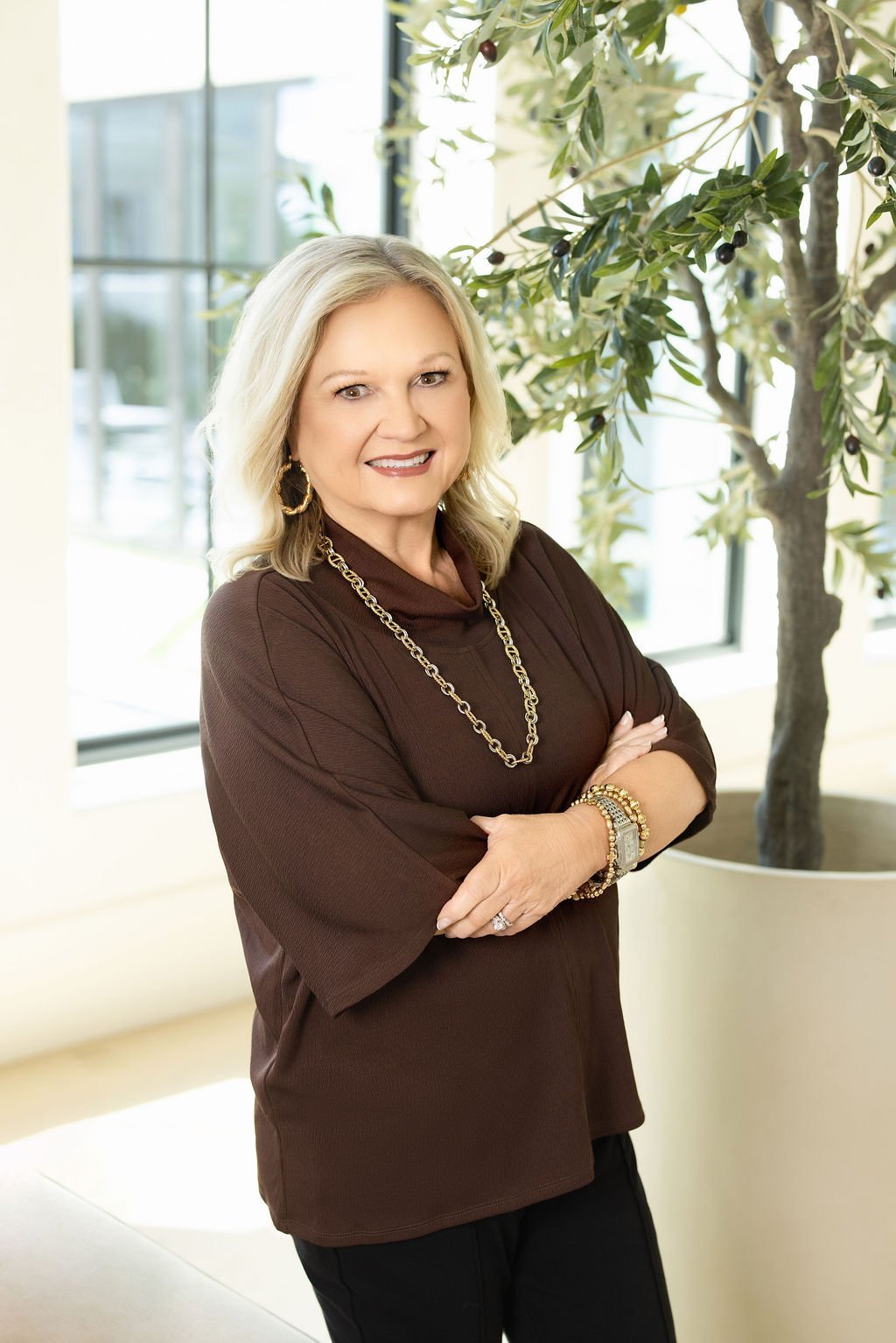 A middle-aged woman wanted updated headshots with blond hair standing in front of a large window with a potted olive tree, wearing a brown top accessorized with gold jewelry, smiling with arms crossed.