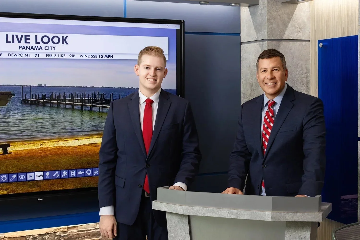 Two men wanting updated professional headshots in suits standing in front of a large screen displaying a live weather update from Panama City. The man on the left is young with light hair, and the man on the right has dark hair. Both are smiling.