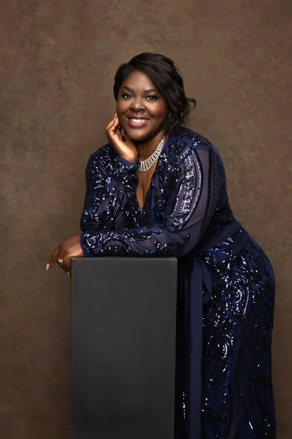 A woman in an elegant navy blue sequined dress poses for a portrait, smiling with her hand resting on her cheek and leaning on a black pedestal, against a plain brown background.