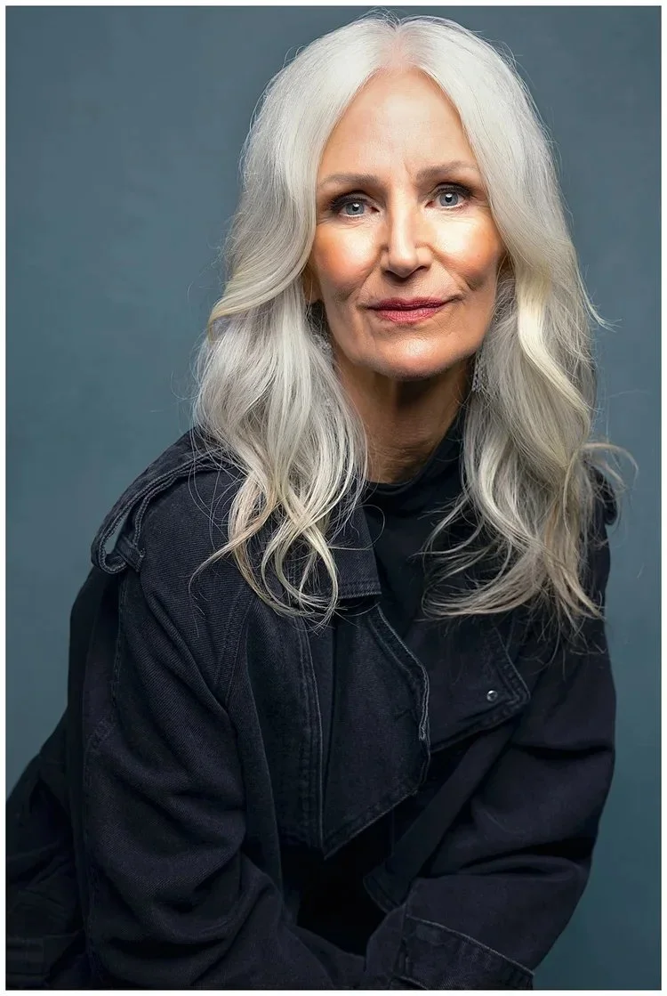 Portrait of an older woman with long, wavy gray hair, wearing a black jacket against a gray background.