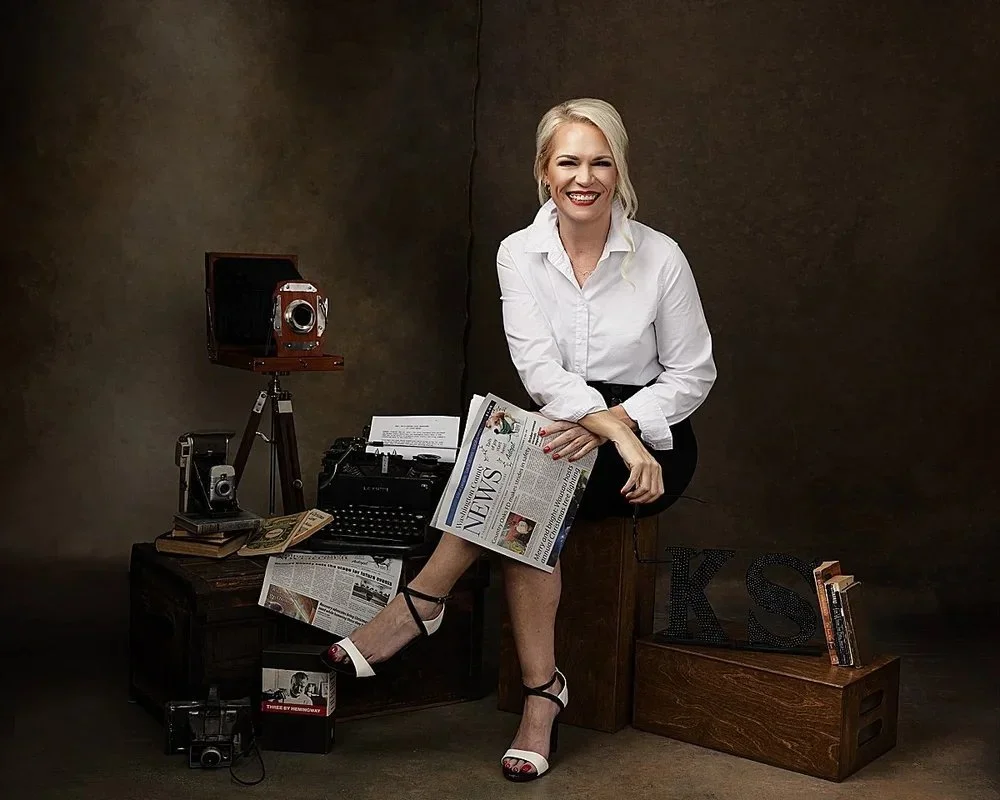 A woman with blonde hair, wearing a white shirt and black skirt, sitting on a wooden box, smiling and holding a newspaper. The background is a dark, textured studio backdrop. There are vintage cameras, books, snacks, and decorative letters around her