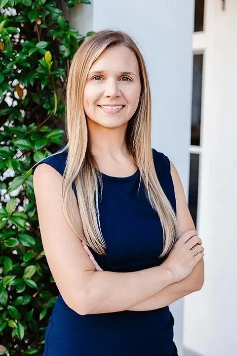 A woman with long blonde hair, wearing a sleeveless navy blue dress, smiling with arms crossed, standing outside in front of green bushes and a modern building.