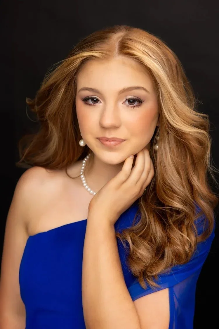 Portrait of a young woman with red wavy hair, wearing pearl jewelry and a blue off-shoulder dress, against a black background.