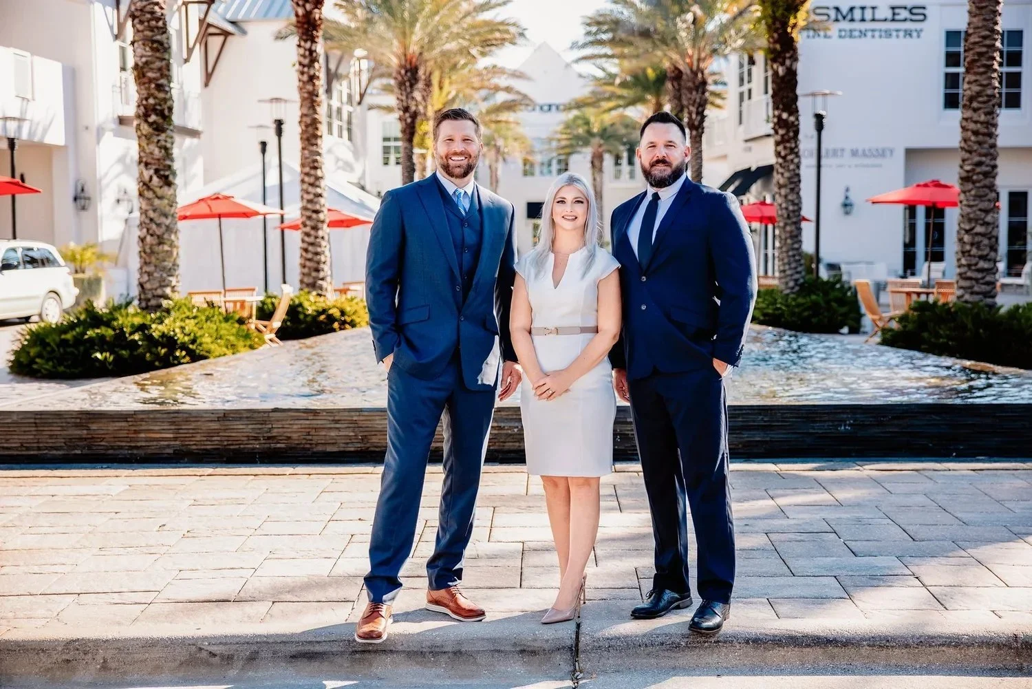 Three professionally dressed individuals, two men and one woman, standing outdoors in a sunny urban area with palm trees, buildings, and outdoor seating with red umbrellas in the background.